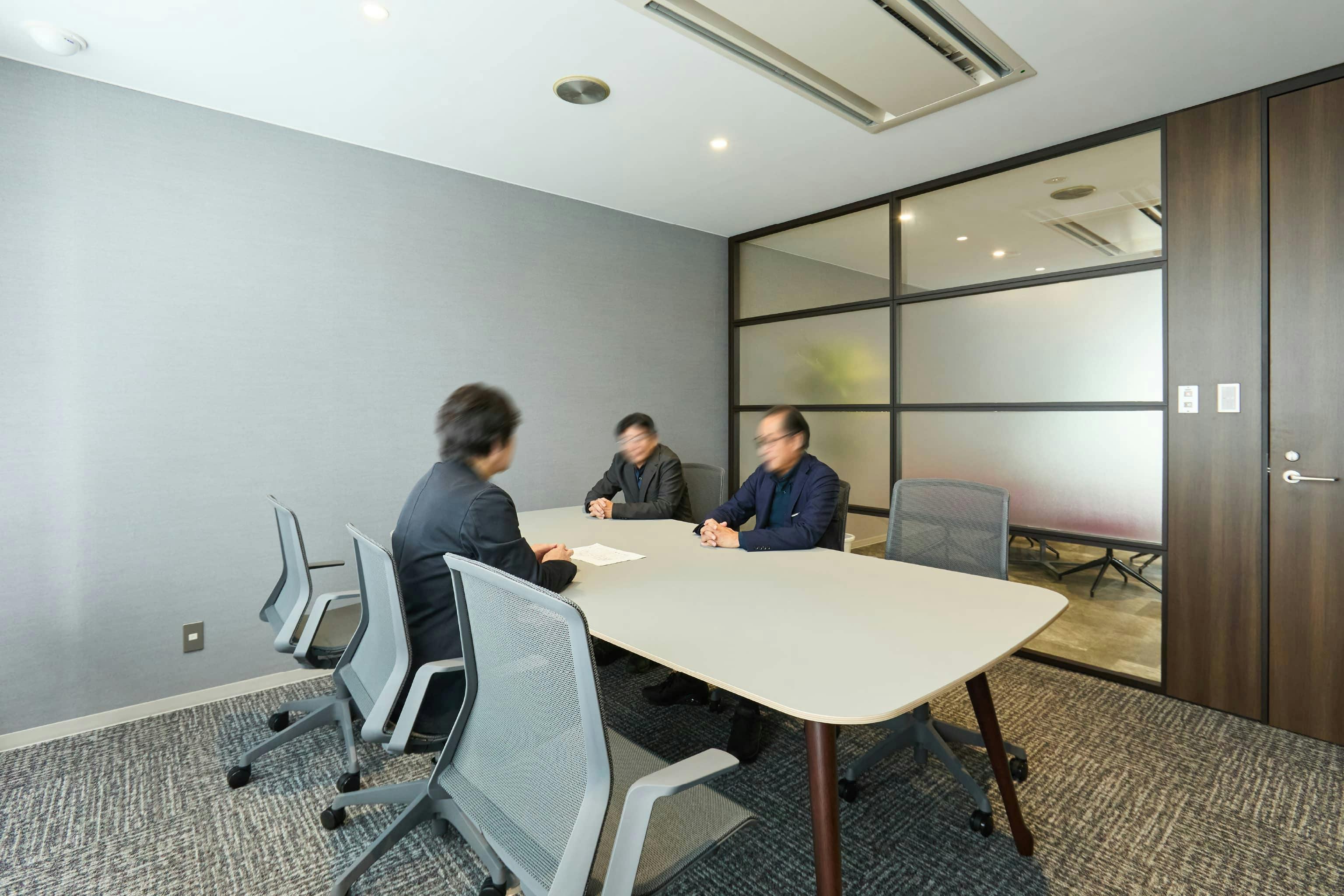 株式会社荒谷建設コンサルタント 様の事例写真／[1F応接室]6人まで利用できる応接室。壁紙やイスの色は、受付との繋がりを意識してグレーで統一されています。一面をガラス壁にすることで、利用状況が外からすぐ分かります。