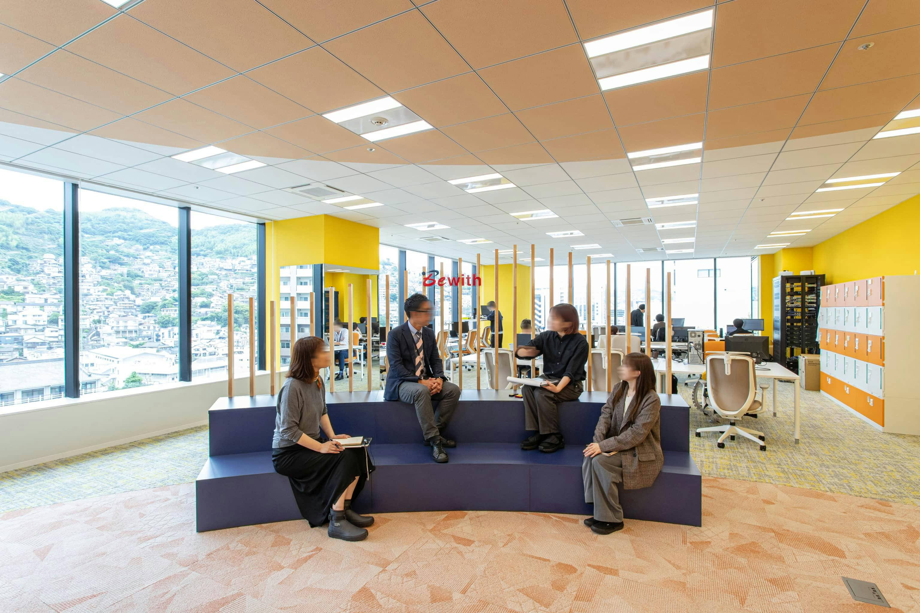 ビーウィズ株式会社 様の事例写真／[共創エリア]天井と床にオレンジの円形モチーフが施され、隣接する執務エリアとの空間をやわらかく区切っています。パーティションを兼ねた造作ベンチはルーバー部分に企業ロゴを配し、抜け感を保ちつつアイキャッチの役割も持たせています。