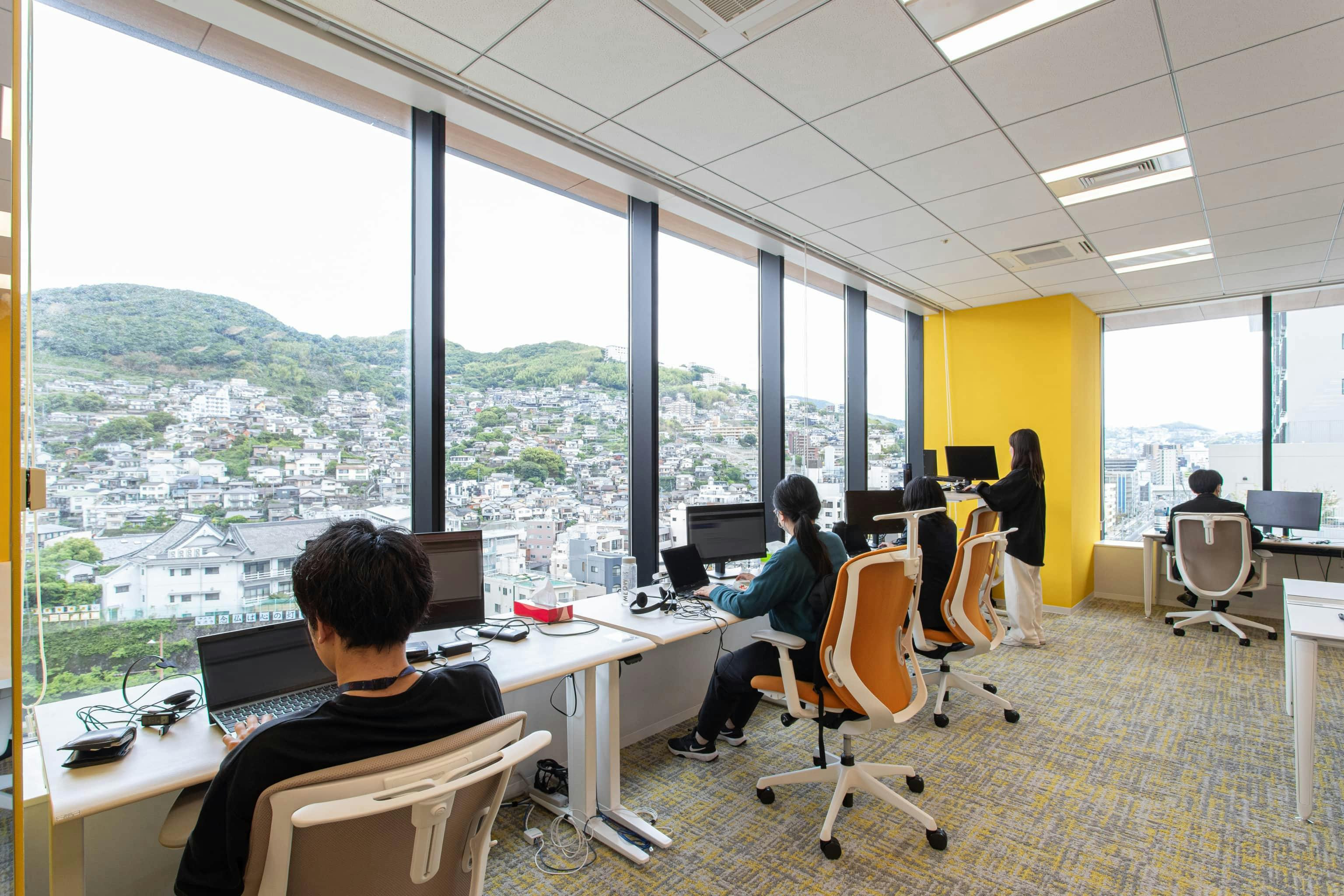 ビーウィズ株式会社 様の事例写真／[執務エリア]電動昇降デスクを配置した窓際席。各自のコンディションに合わせて１日の中でデスクの天板を上げたり下げたりしながら、業務に取り組むことが可能です。眺望も素晴らしく、人気のスペースとなっています。