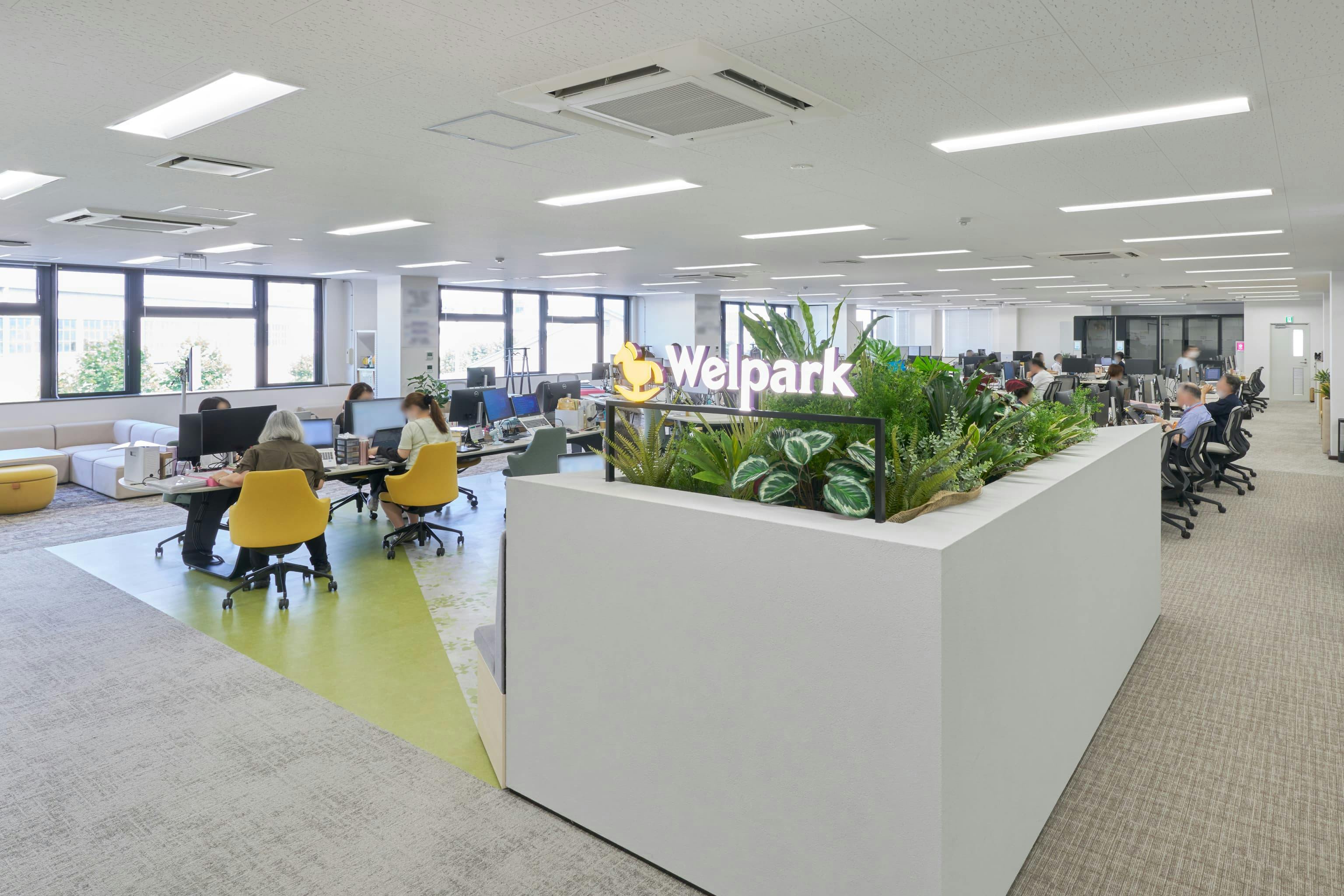 株式会社ウェルパーク 様の事例写真／[エントランス付近のサイン]エントランス付近には、アイキャッチとなる社名サインとともに、植栽をあしらった什器を配置。セキュリティに配慮し、来訪者がフロアに入った際に執務エリアを奥まで見通せないよう、さりげなく目隠しをしています。