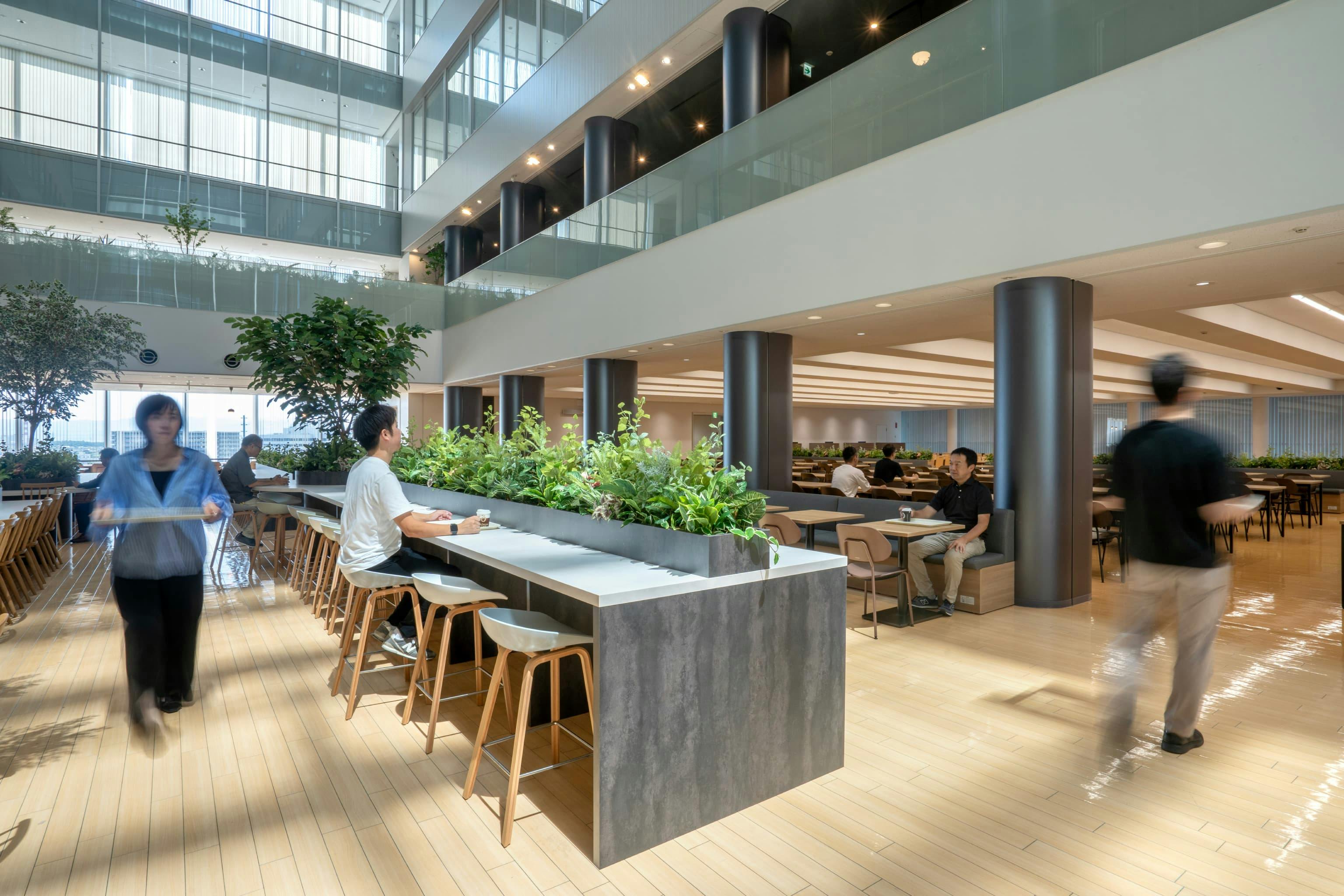株式会社村田製作所 様の事例写真／[5階食堂（アトリウム）]手早く食事を済ませたい人向けのハイカウンター席。配膳エリアに近い席の回転数を上げることで、食堂全体の混雑を緩和しています。卓上のグリーンは適度な目隠しとなり、つかの間の癒やし効果をもたらします。