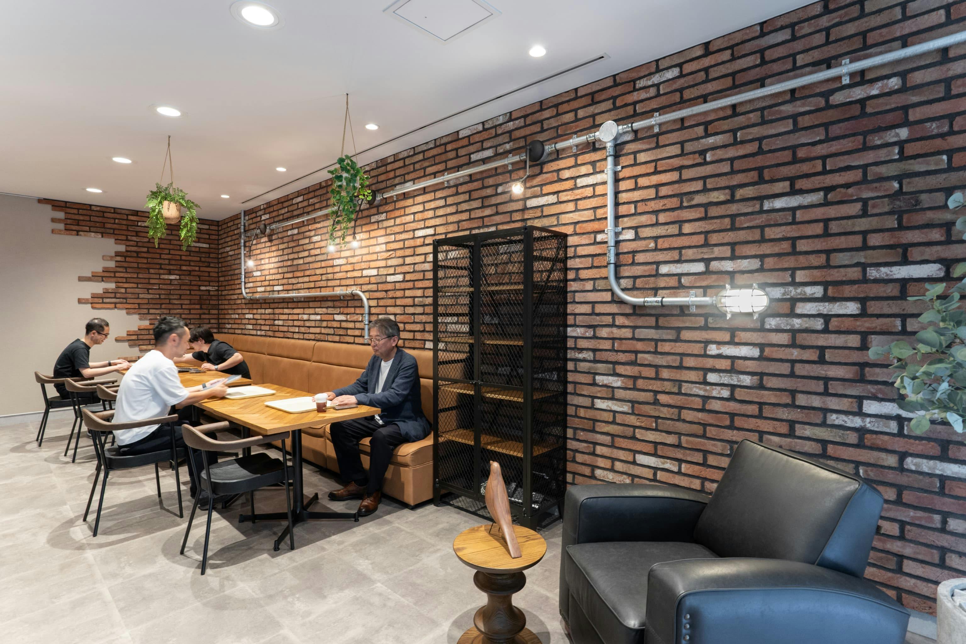 株式会社村田製作所 様の事例写真／[5階食堂会議室]レンガの質感を活かした壁紙に本物のパイプがあしらわれ、カジュアルながら落ち着いた雰囲気。ヘリンボーン天板のテーブルが置かれた革張りのソファ席に座れば、カフェに居るような気分で会話が弾みます。