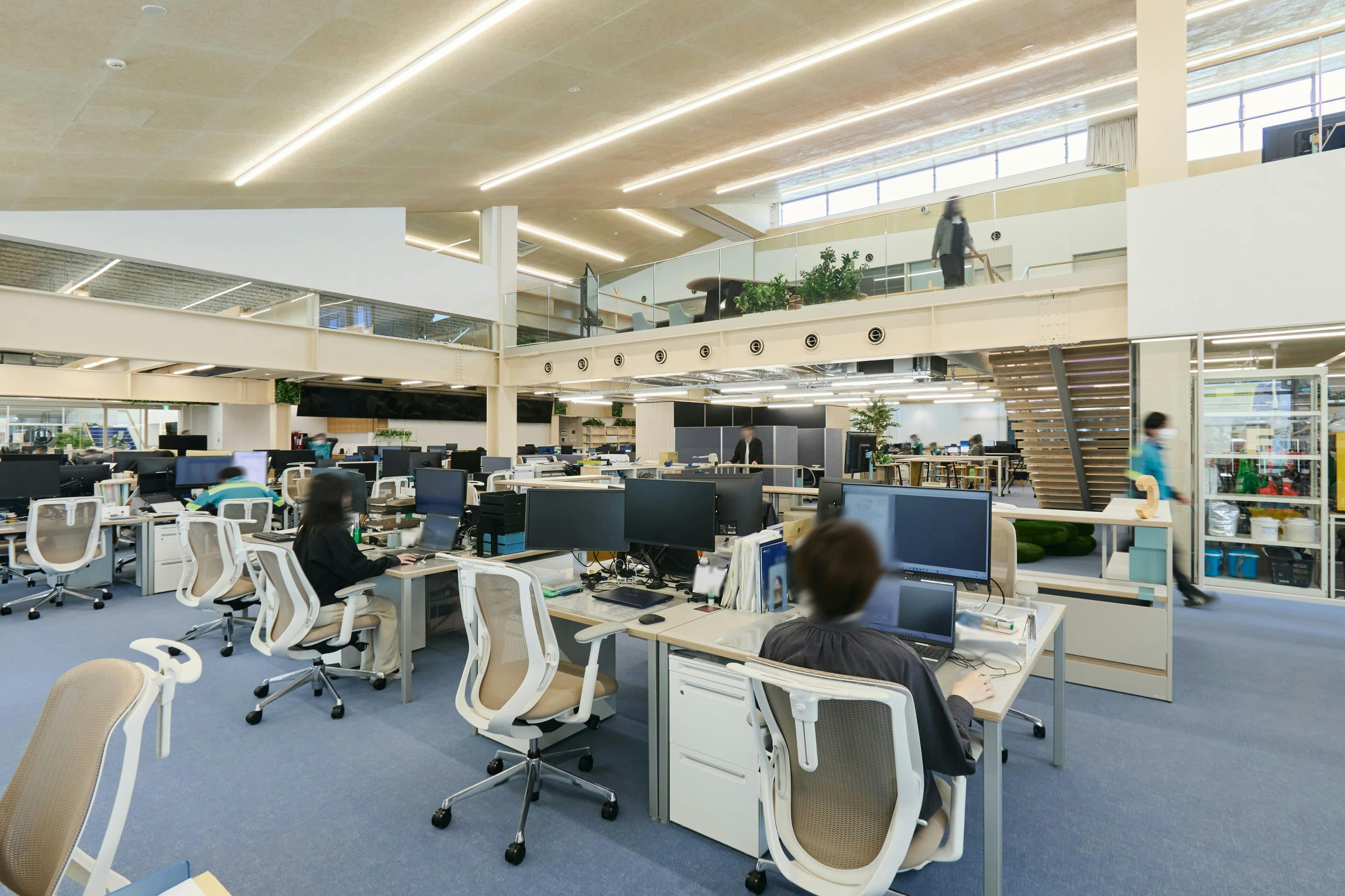 今治造船株式会社 様の事例写真／[1F執務エリア（オフィス棟）]デスクは柔軟にレイアウト変更ができるスクエアタイプを採用することで、将来的な増席や組織変更にもスムーズに対応可能です。執務エリアからは中2階フロアの様子がうかがえ、大きな吹き抜けが開放感を与えています。