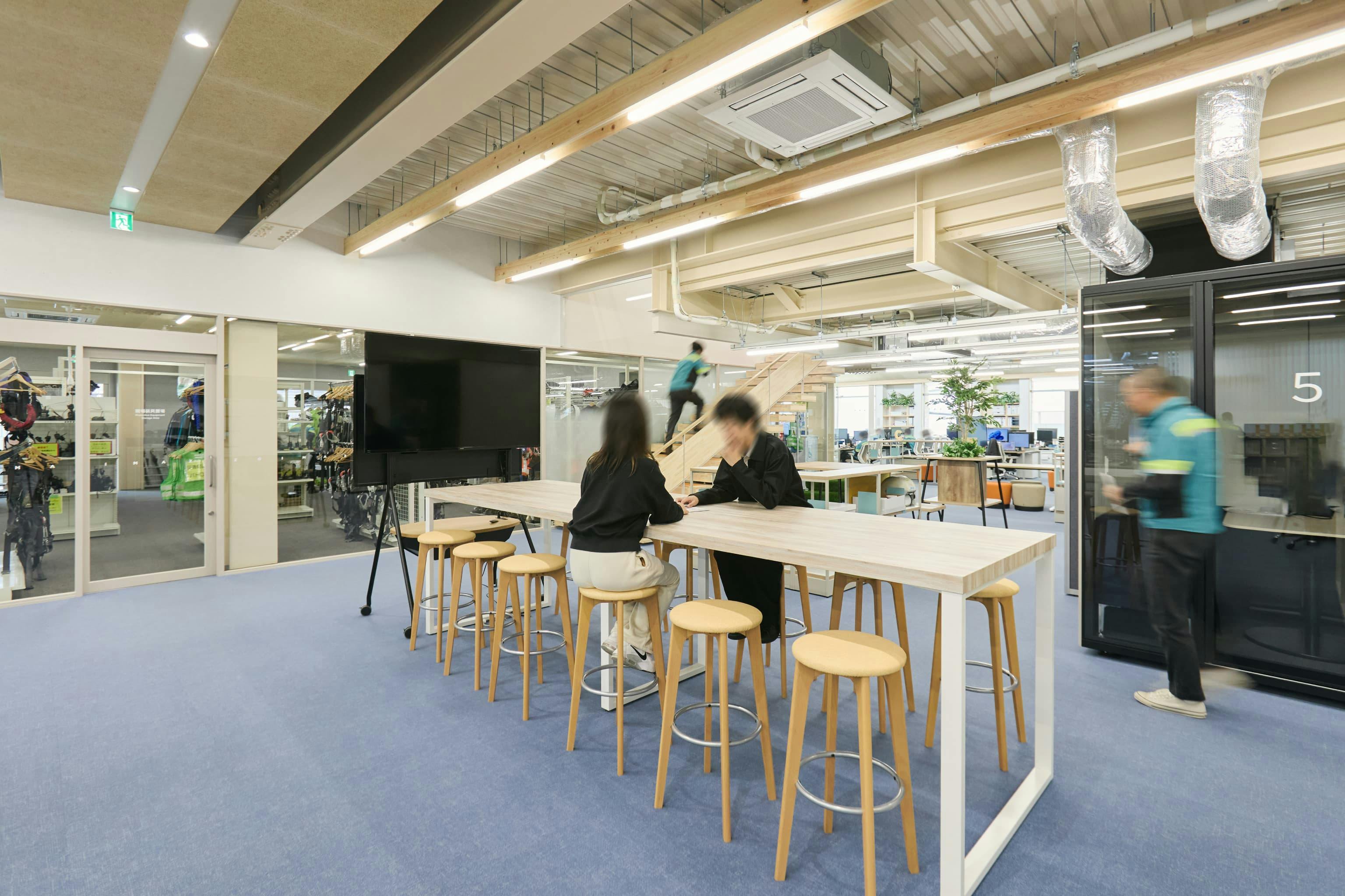 今治造船株式会社 様の事例写真／[1F交流エリア（オフィス棟）]交流エリアに配置された大型ハイテーブルは、チーム単位での利用にも便利。さっと座れるハイスツールを置くことで、立ち寄りやすく、さらに歩いている人と同じ目線の高さになり話しかけやすくなっています。