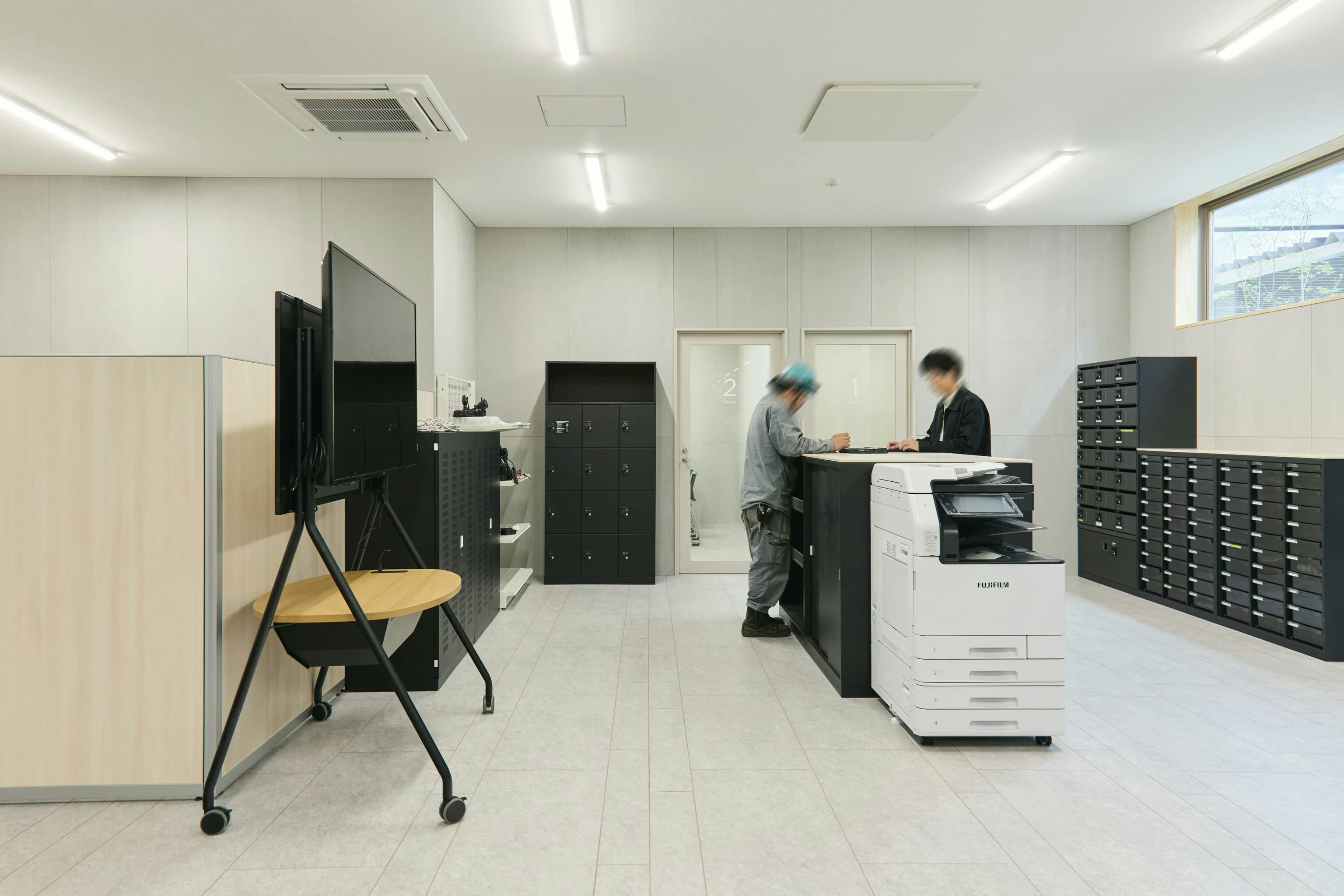 今治造船株式会社 様の事例写真／[1F協力会社との打ち合わせスペース（オフィス棟）]協力会社との打ち合わせスペースでは、立ち話やちょっとした打ち合わせも可能な場が備わっています。図面や資料を出力・確認するためのコピー機や作業台、ディスプレイが置かれ、打ち合わせを効率的に進められます。