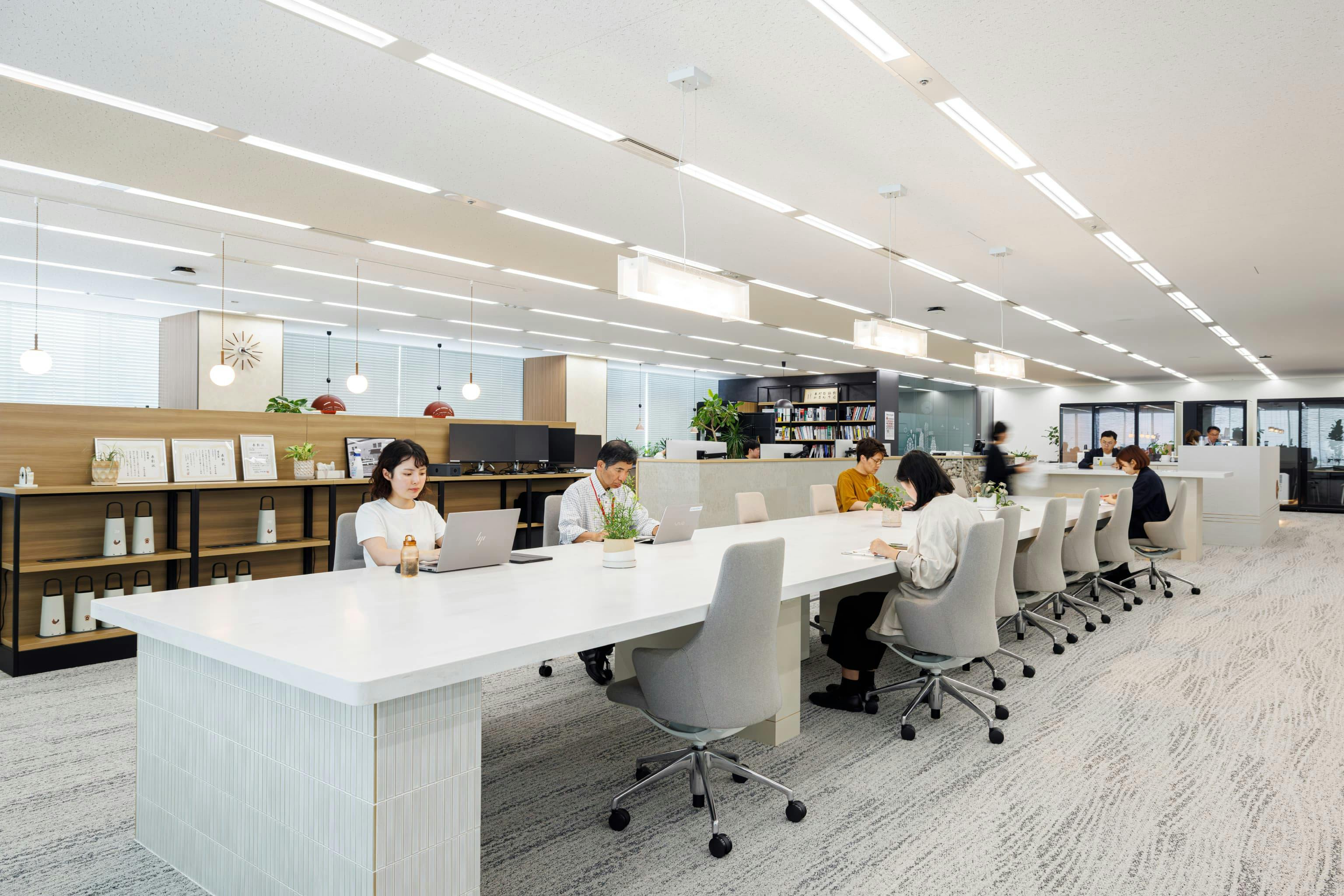 株式会社オカムラ 名古屋ラボオフィス『Cent Labo』の事例写真／[ランウェイエリア]白を基調とした「ランウェイ」に対して、写真後方の執務エリアは落ち着いた茶色をベースとし、色調の違いでゾーニング。人工大理石の長テーブルは、タッチダウンオフィスとして他拠点の社員が利用することも。