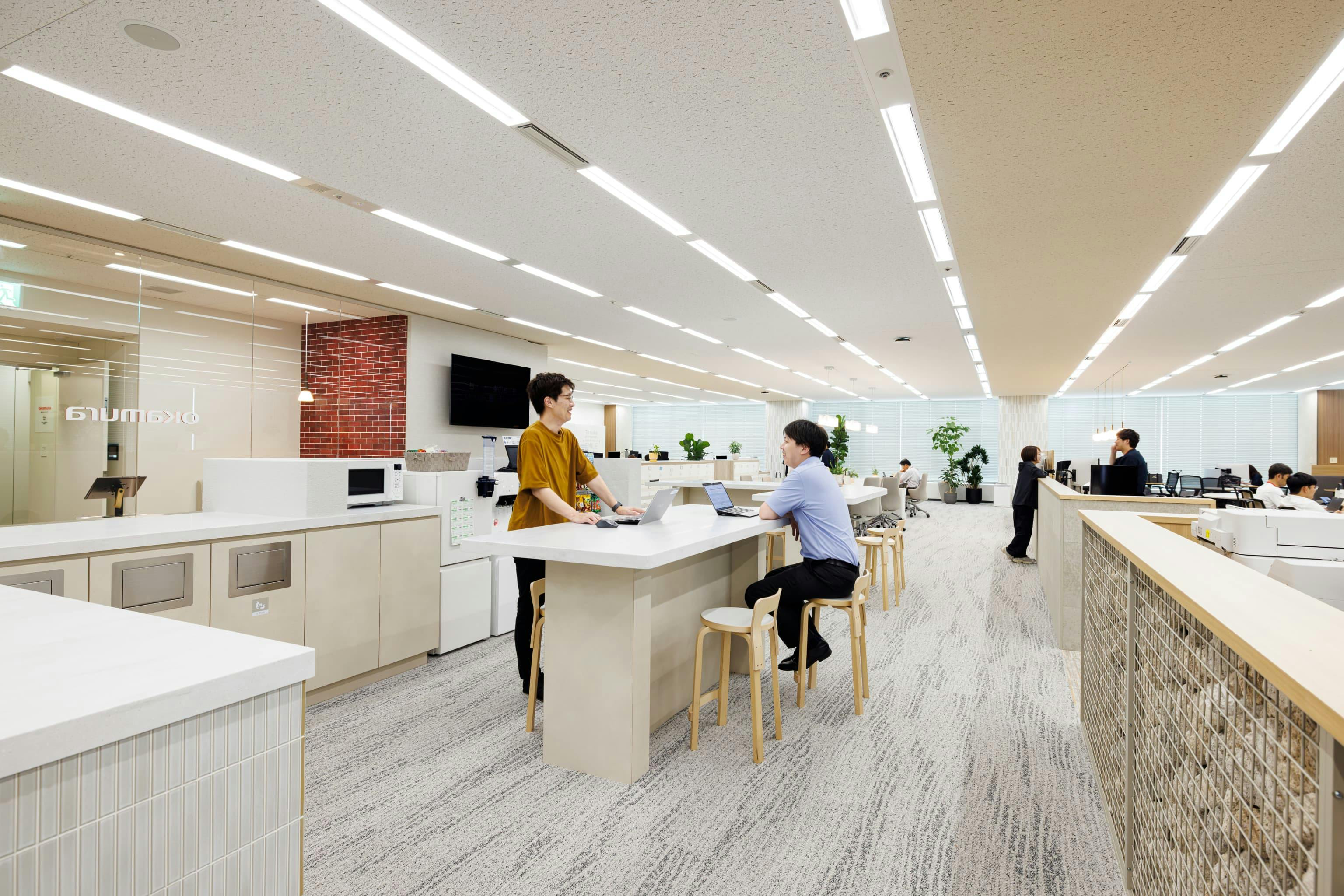 株式会社オカムラ 名古屋ラボオフィス『Cent Labo』の事例写真／[カフェスペース（ランウェイエリア内）]造作のハイカウンターを設置し、カフェスペースの機能を持たせています。人通りが多く、雑談しやすいスポットです。飲み物を手にラフな打ち合わせが始まったり、すき間時間に立ち作業をしたりする姿が見られます。