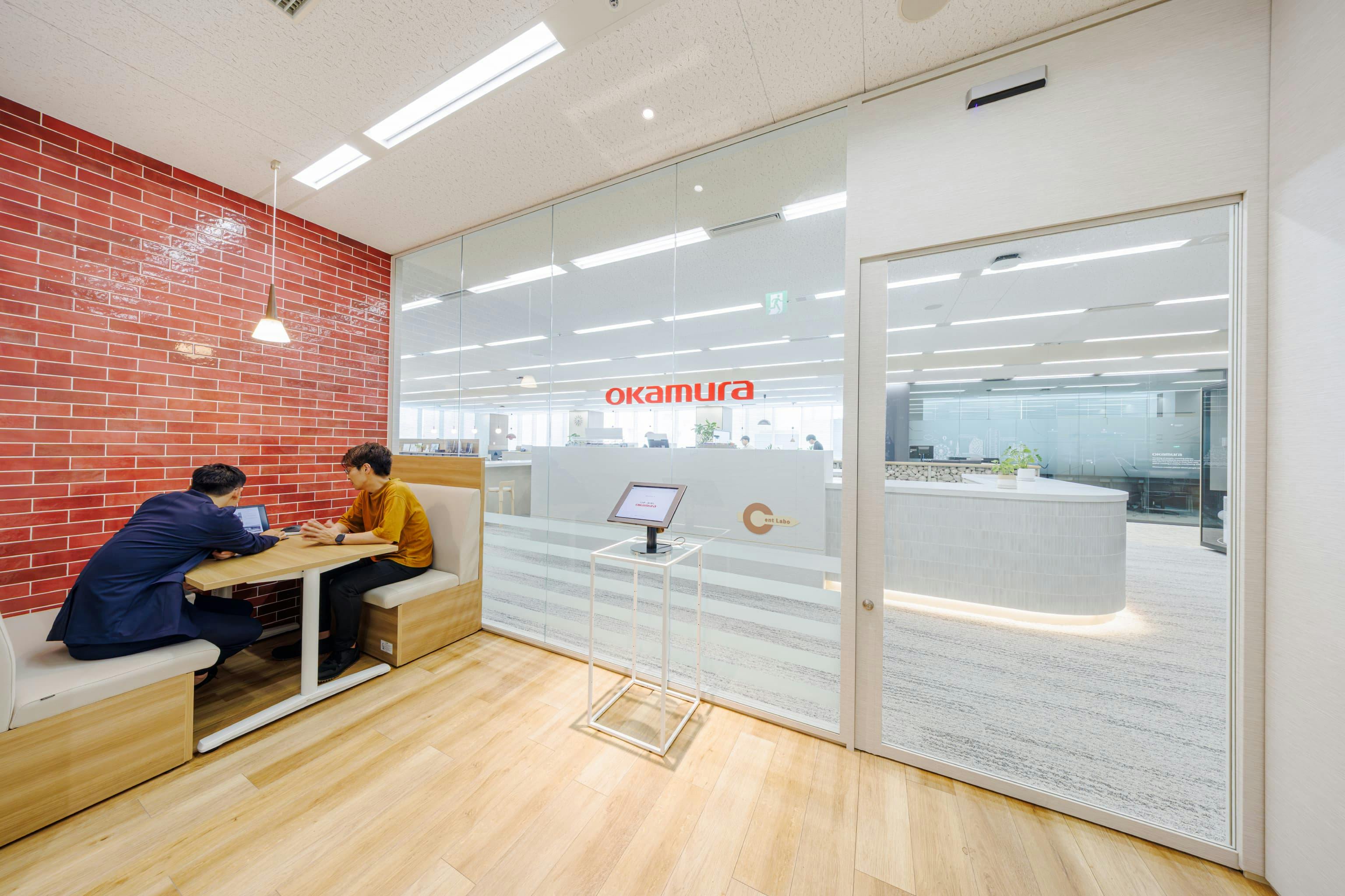 株式会社オカムラ 名古屋ラボオフィス『Cent Labo』の事例写真／[エントランスの打ち合わせスペース]エントランス外の一角にファミレス席を設け、待ち合わせや社外打ち合わせに活用。壁面にはコーポレートカラーの赤いタイルが貼られています。エントランスはガラス面を大きく設け、広がりのある空間に。