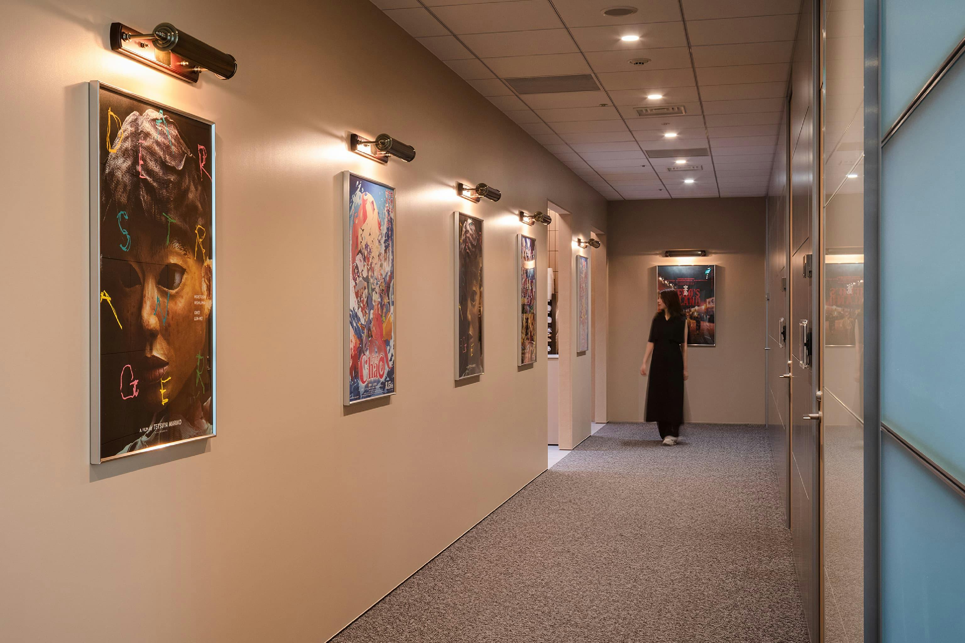 東映株式会社 様の事例写真／[9階 廊下]試写室や応接室、会議室に隣接する廊下にはポスターが掲示され、映画館さながらの雰囲気に。居抜きオフィスの壁やカーペットをそのまま活かしつつ、ポスターを照らす照明が公開待機作品の世界に触れられる空間をつくり出しています。