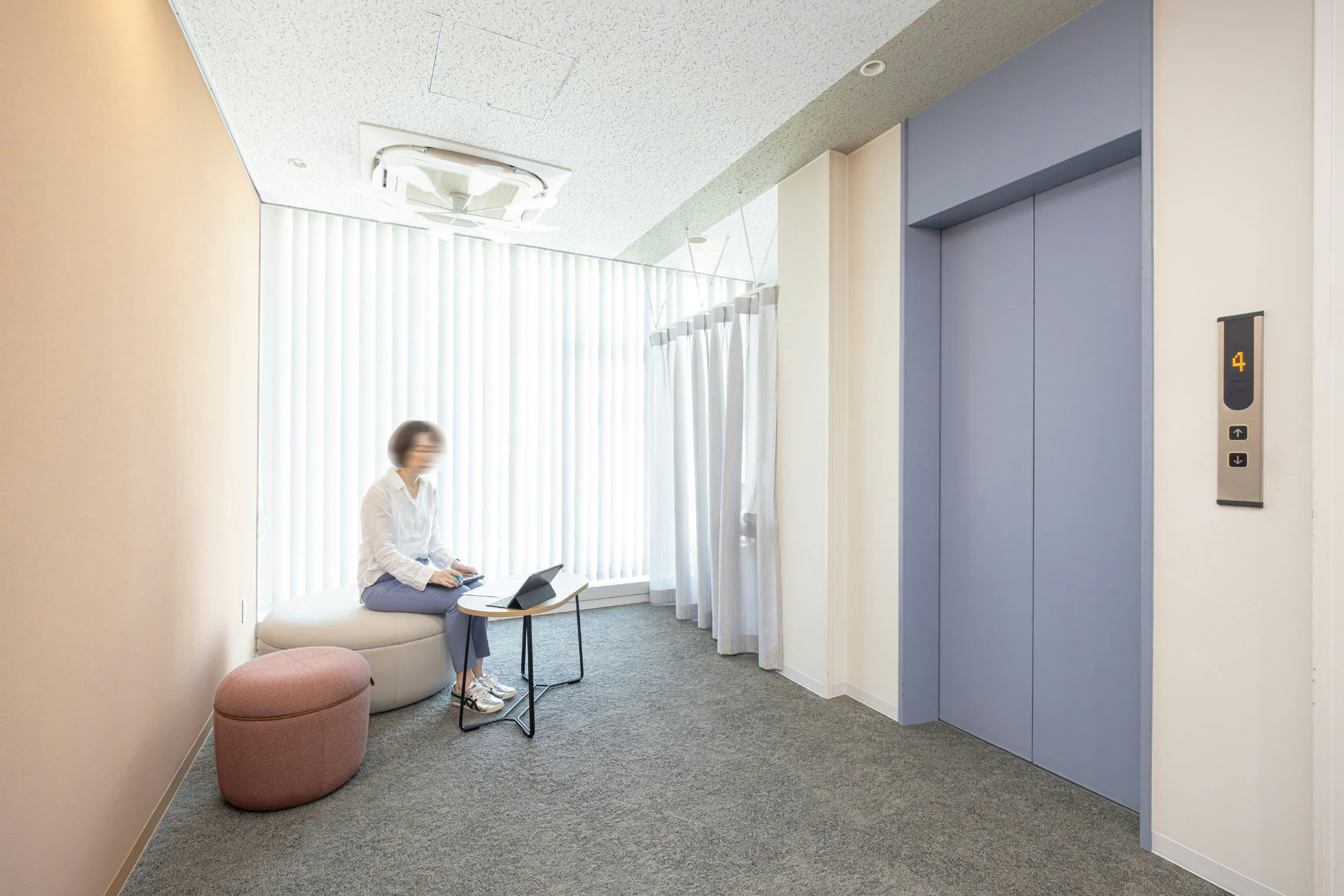 日本ガスケット株式会社 様の事例写真／[エントランス]エレベーター前には、待ち合いスペースが用意されています。暖色の壁と呼応する温かな色味のチェアが来訪者の心を和ませます。