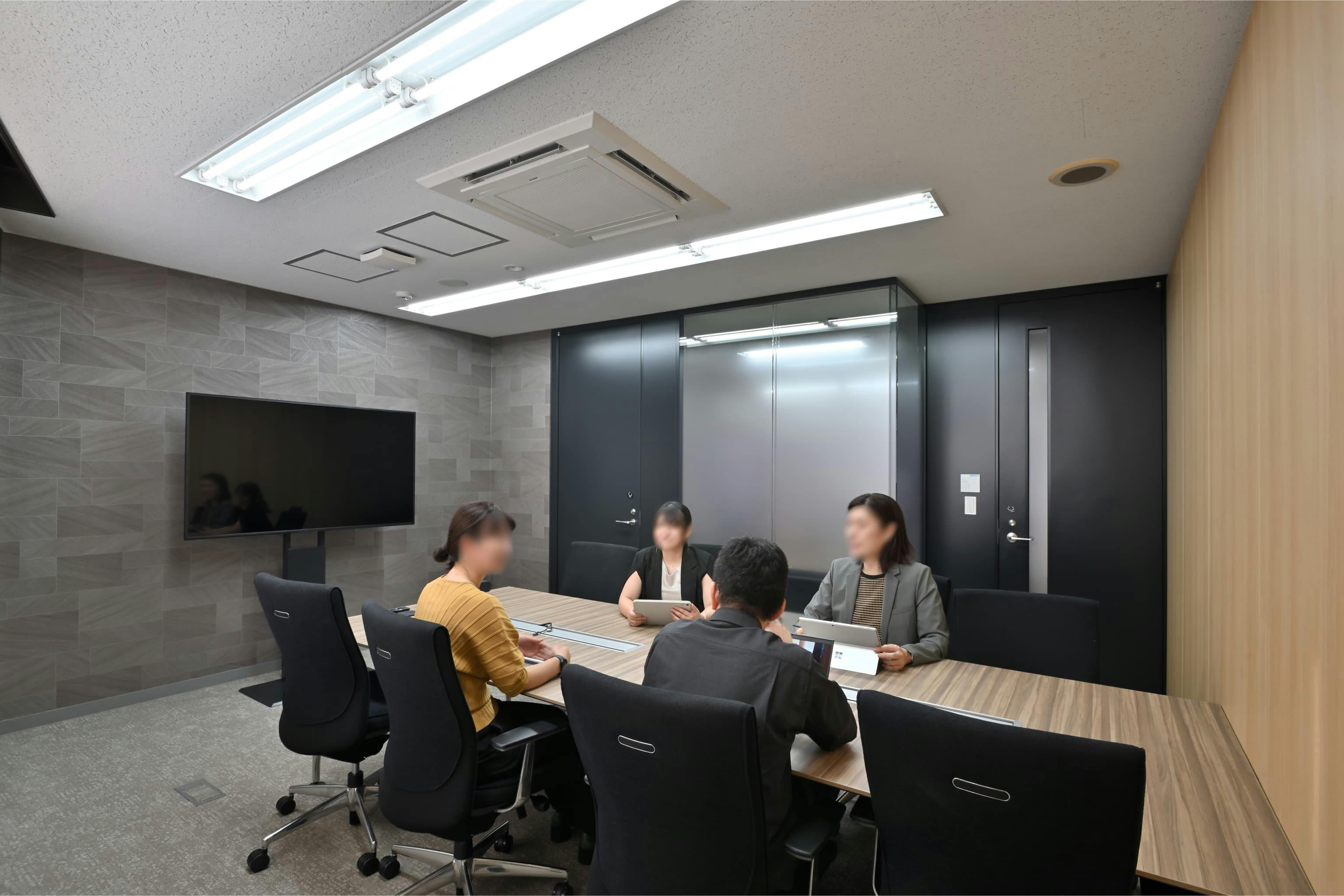 株式会社エスケイケイ 様の事例写真／[応接室]モノトーンの什器とグレーのタイル壁が、重厚な雰囲気をつくっています。エントランスに隣接し、執務エリアを通らずに来訪者を案内できます。落ち着いた設えの中で、商談や会議に集中できる空間です。