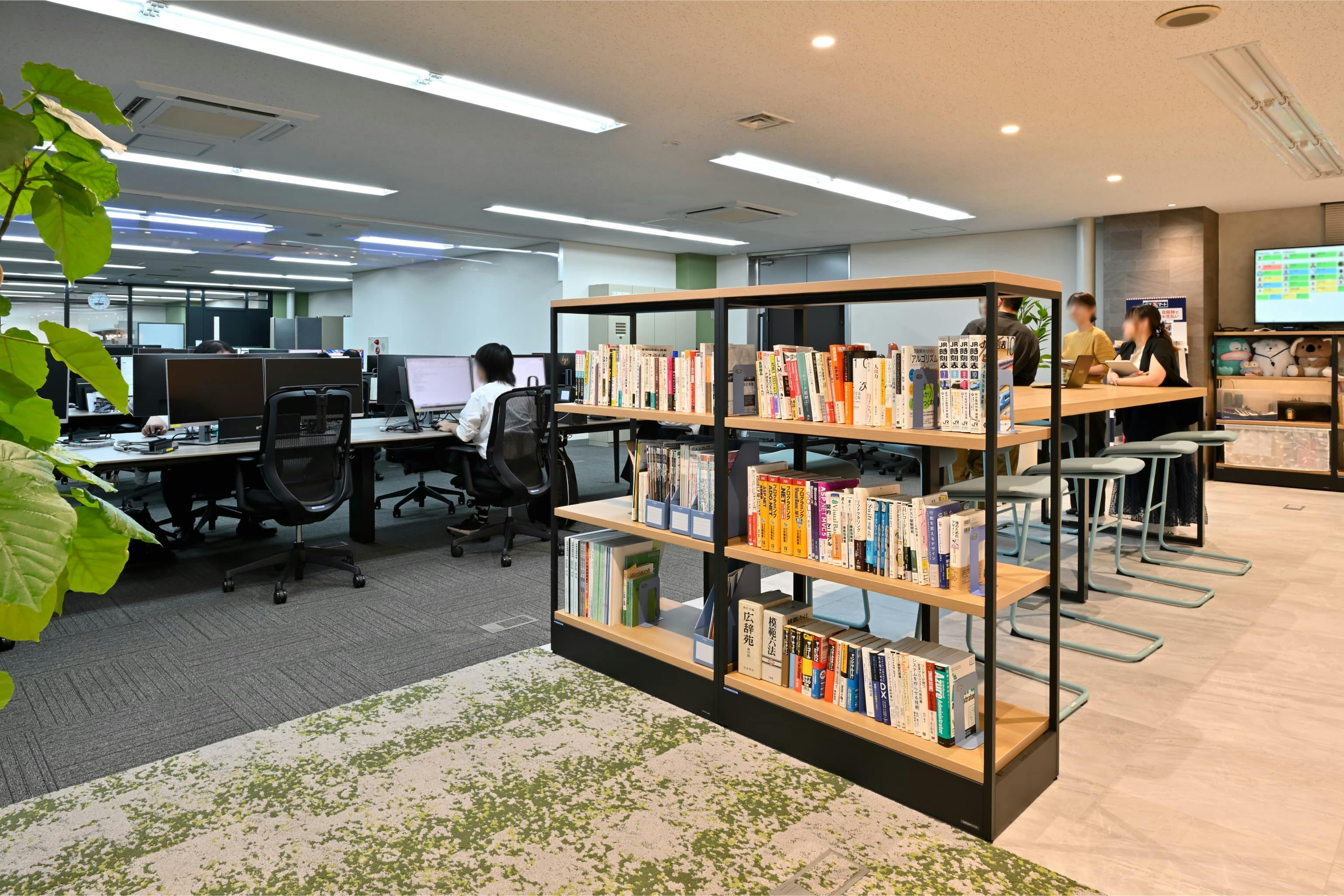 株式会社エスケイケイ 様の事例写真／[交流エリア]本棚には実用書が並び、気になったことをその場で調べられるちょっとした学びの場にも。書籍をきっかけに話題が広がり、仕事のヒントを共有し合う光景も見られます。
