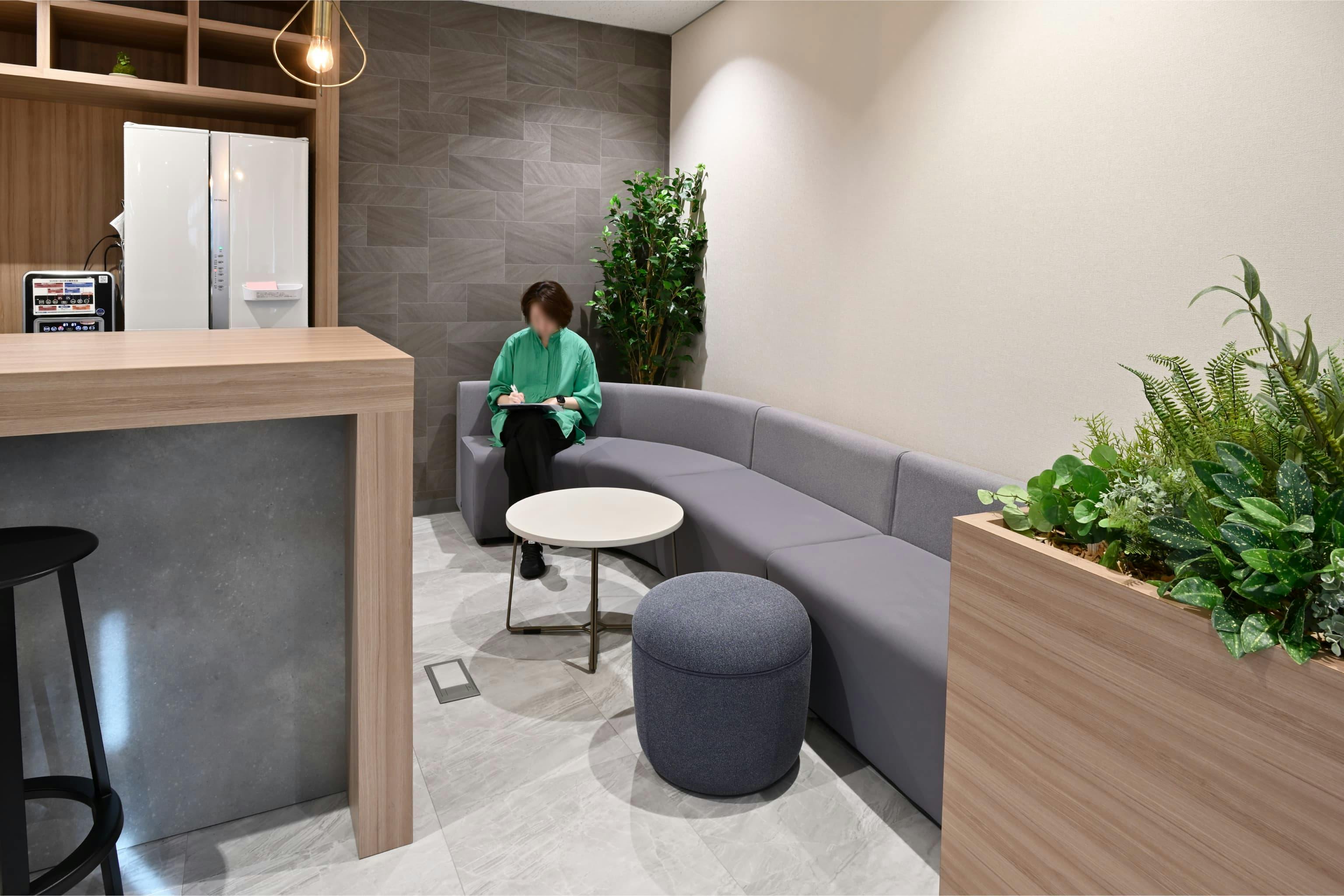 株式会社エスケイケイ 様の事例写真／[交流エリア（カフェスペース）]カウンターの横にある小さなコーナーは、ほどよく囲われる安心感で落ち着ける場所。丸いテーブルと曲線ソファがやわらかな雰囲気をつくり、お昼を食べたり、おしゃべりしたりと、そっと気持ちをほぐしてくれます。