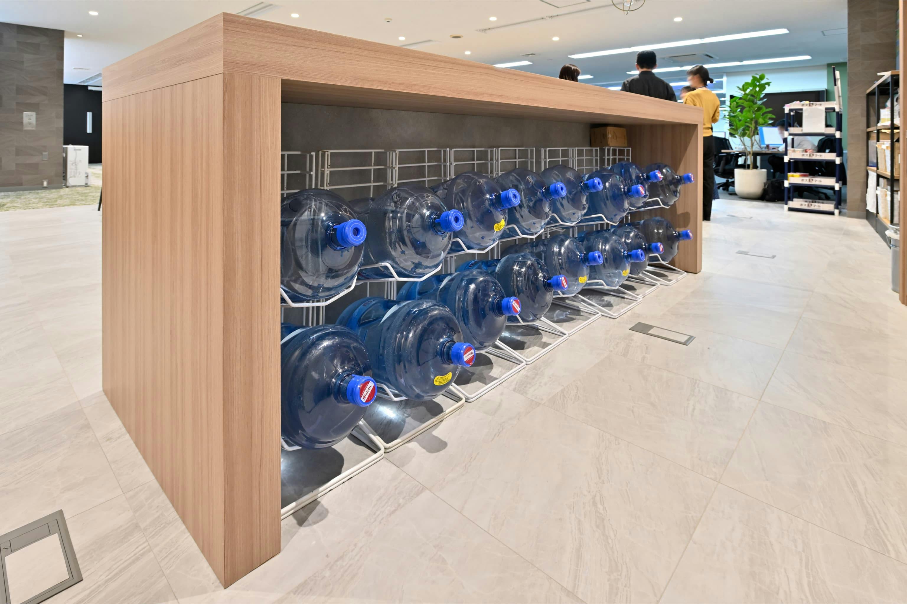 株式会社エスケイケイ 様の事例写真／[交流エリア（カフェスペース）]カウンター下のスペースを、ウォーターサーバーボトルのストック置き場として活用。必要量をすっきり収められるように寸法を整えて造作されており、デッドスペースが機能的な収納場所として生かされています。