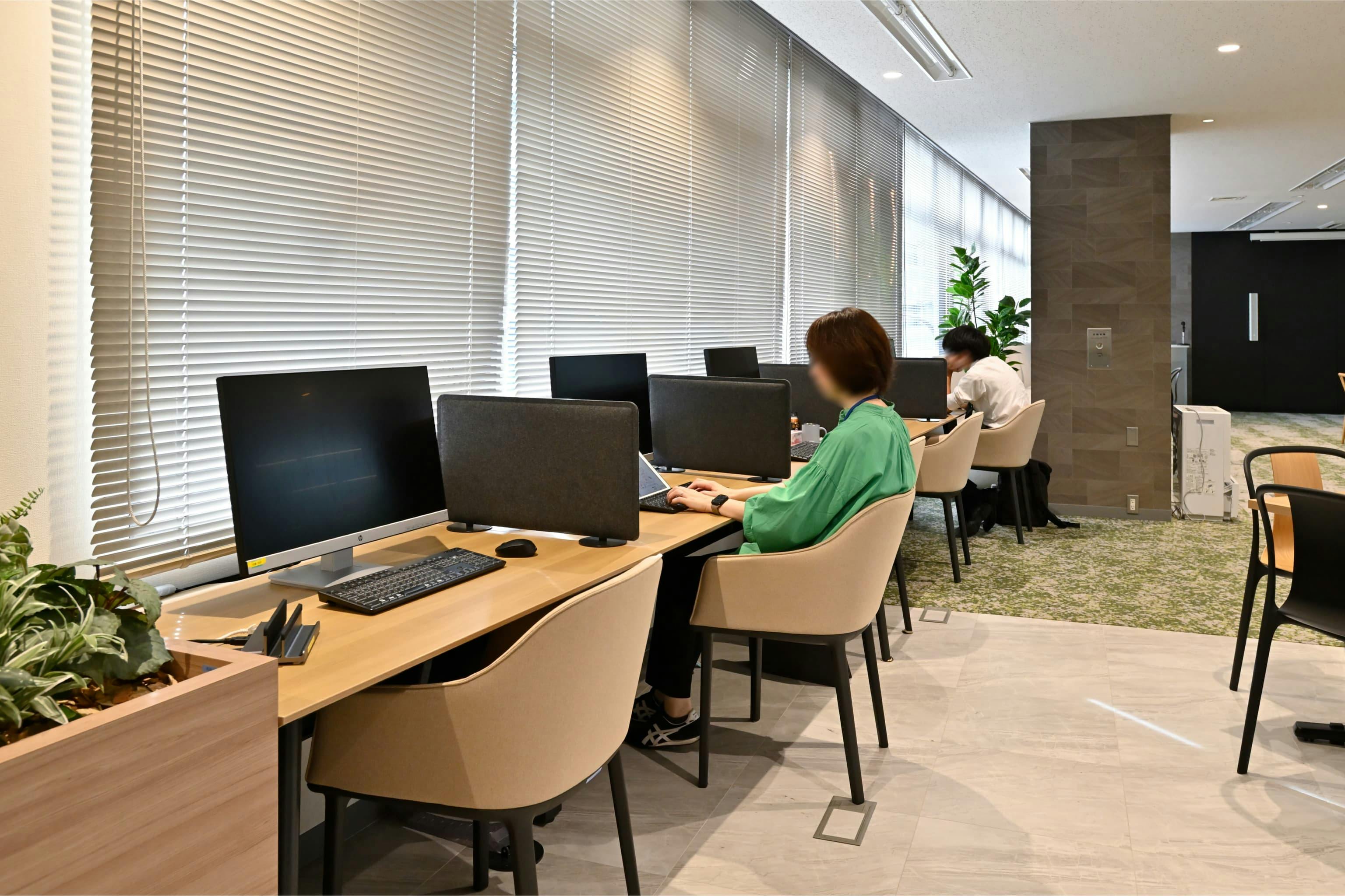 株式会社エスケイケイ 様の事例写真／[交流エリア（カフェスペース）]窓際の席では、外の景色を眺めながら気分を切り替えて作業できます。近くで交わされる会話がほのかに耳に届き、まるで街中のカフェにいるような心地よさ。かすかなざわめきが適度な刺激となり、集中力も高まります。
