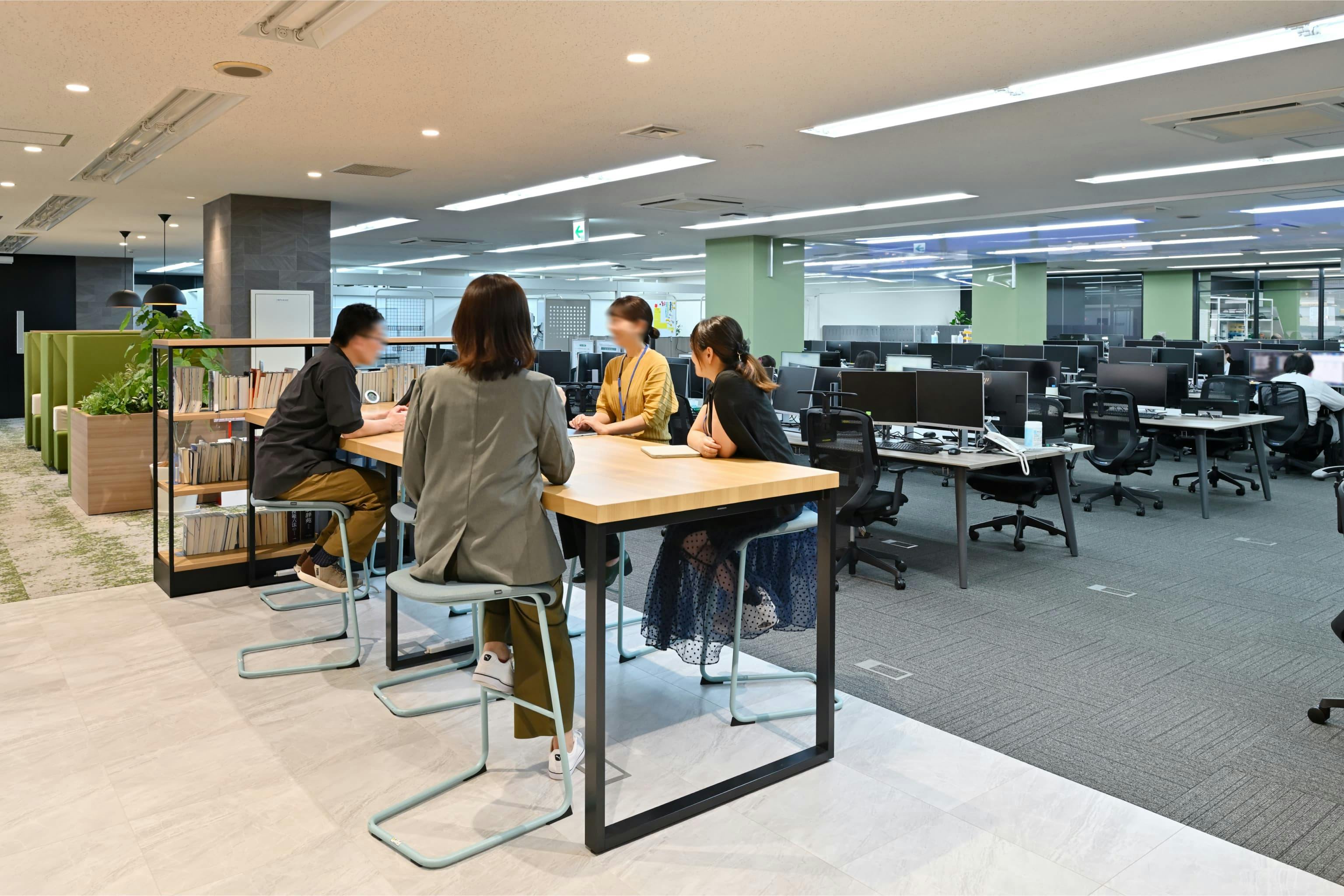 株式会社エスケイケイ 様の事例写真／[交流エリア（カフェスペース）]執務エリアとの境に置かれたハイテーブルは、立ち寄りやすい気軽さが魅力です。ここで交わされる会話は周囲にもほどよく広がり、ゆるやかな情報共有の場にも。立ち話の延長で行うクイックな打ち合わせにもぴったりです。