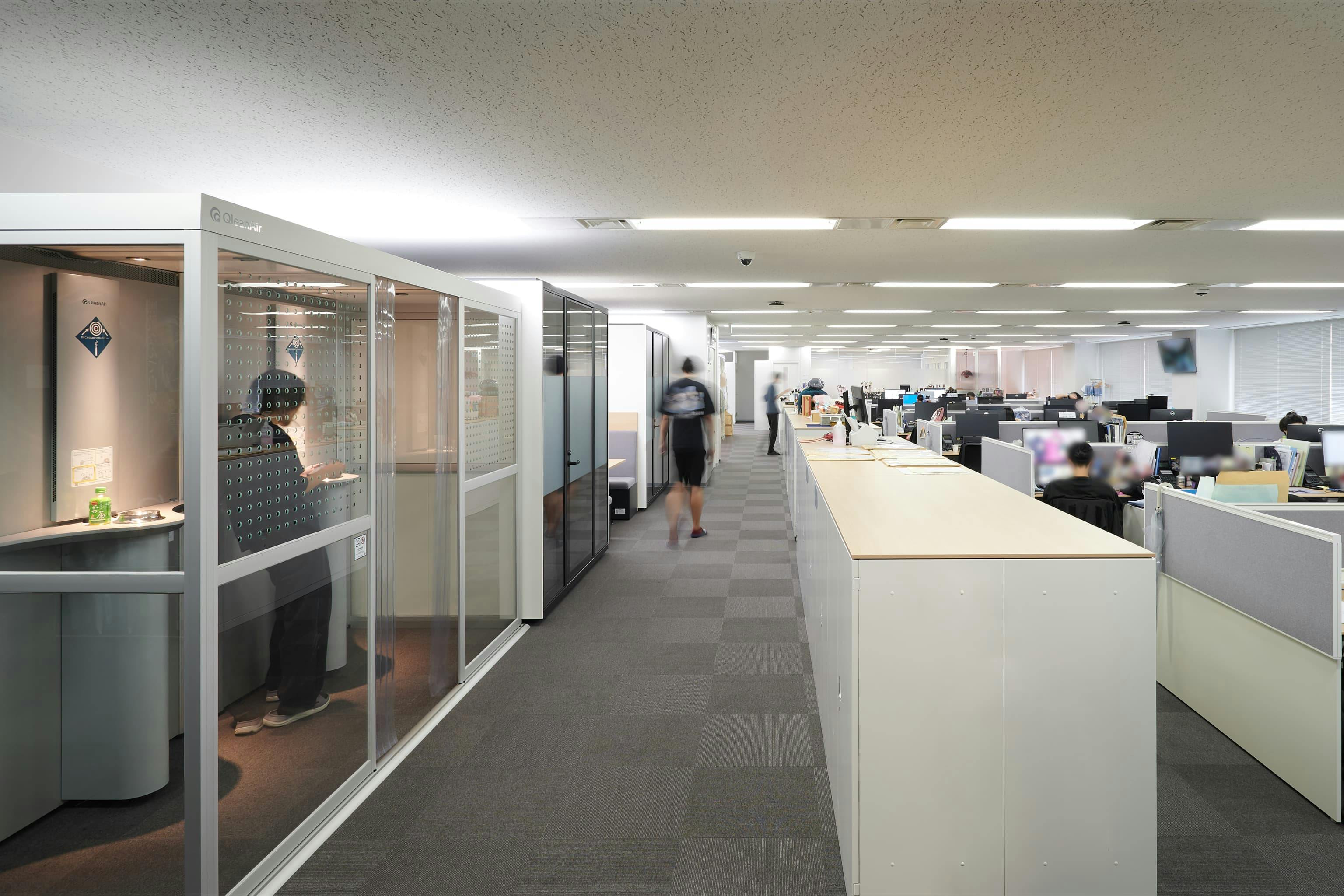 株式会社A-1 Pictures 様の事例写真／[N棟3F執務エリア]通路脇に設置された喫煙ブースは、リラックスした会話や情報交換が生まれる場となっています。非喫煙者にも配慮されたつくりで、扉がなくとも煙やにおいを遮断。内部が見えるクリアな構造で、誰がいるのかひと目で分かります。