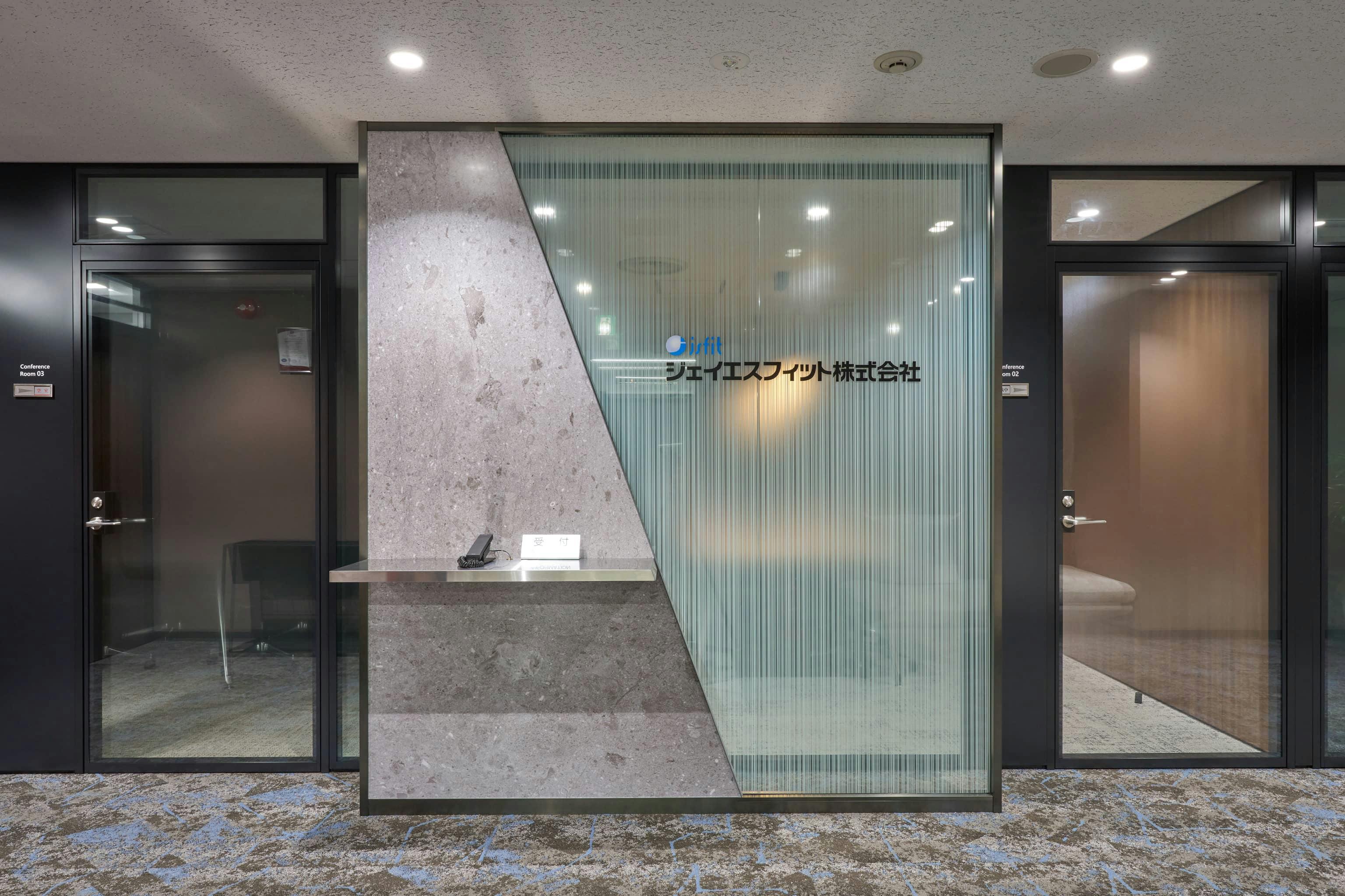 ジェイエスフィット株式会社 様の事例写真／[5Fエントランス]水の流れのようなガラスシートに社名ロゴが重ねられ、立体的に浮かび上がるよう演出。空間に奥行きが生まれています。台形をモチーフにした石目調のデザインがアクセントとなり、凛とした空気感が醸し出されています。