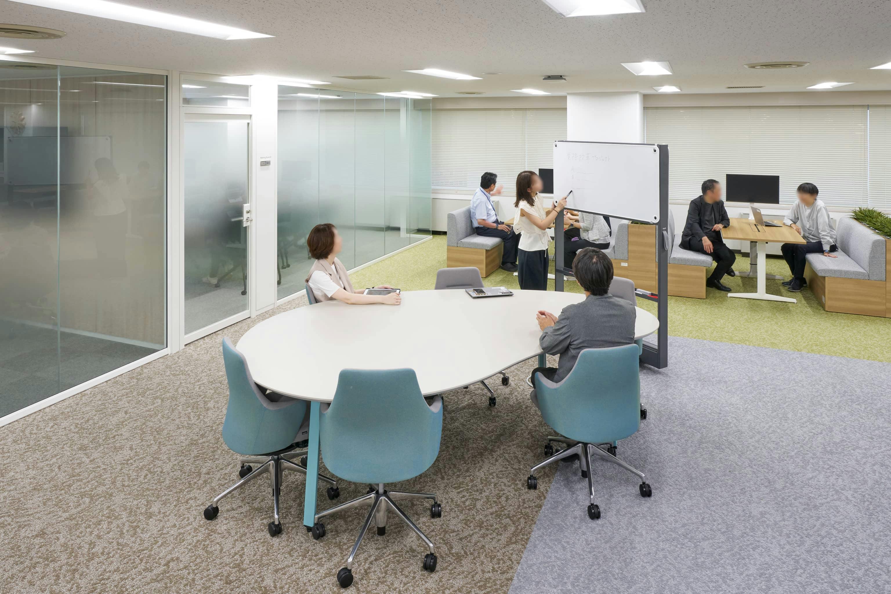 ジェイエスフィット株式会社 様の事例写真／[7F ABWエリア]曲線を描いたビーンズ型のテーブルは、適度な距離感が心地よく、自然体で会話が進みます。通路沿いのため、通りがかった人に軽く意見を求めたり雑談したりと、偶発的なコミュニケーションも生まれます。