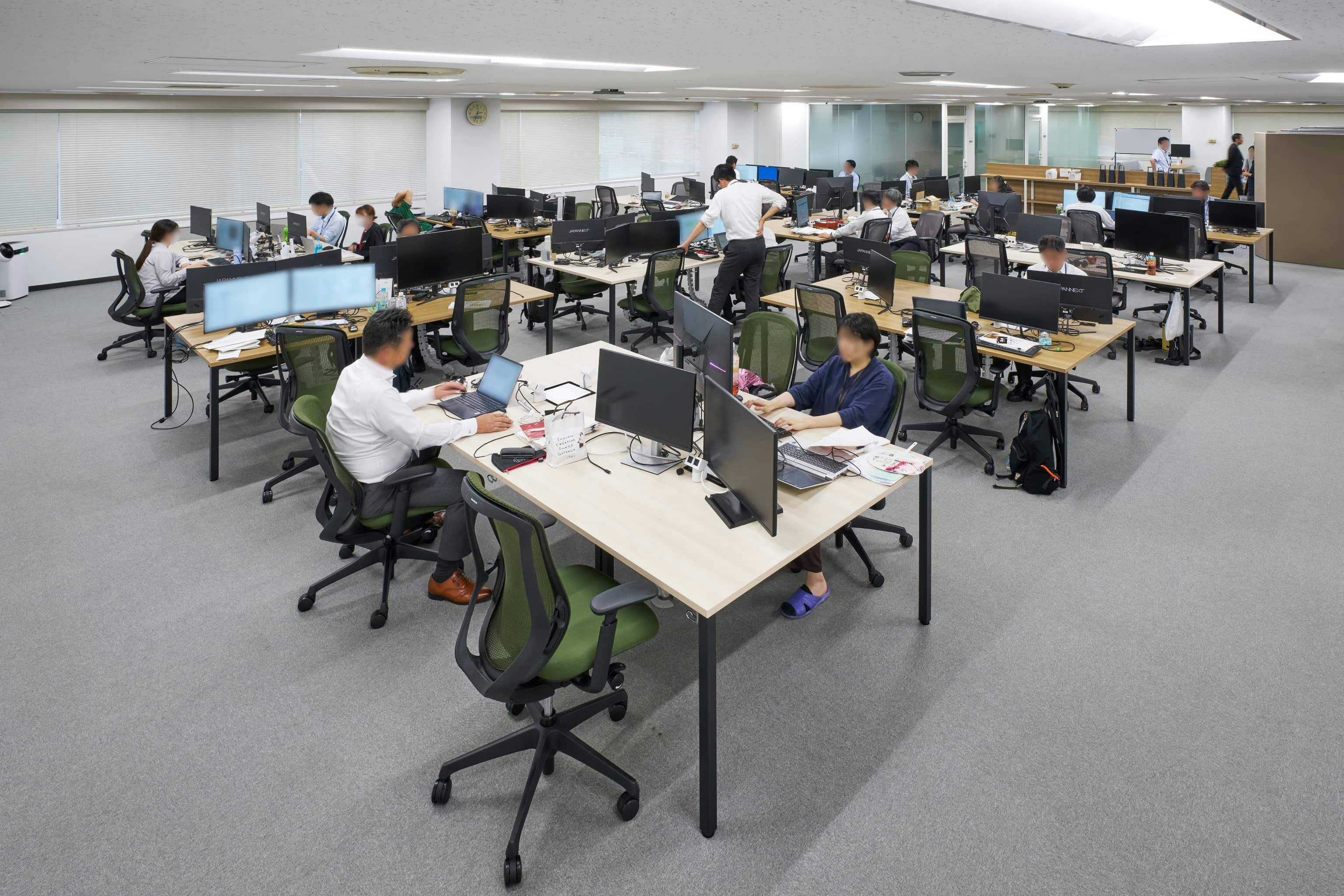 ジェイエスフィット株式会社 様の事例写真／[7F執務エリア]デスクを縦横に組み合わせた変則的な島型レイアウトが動線を自然に交差させ、偶発的なコミュニケーションを生み出します。細身の脚を持つデスクのため視線が抜け、空間全体がすっきりした印象です。