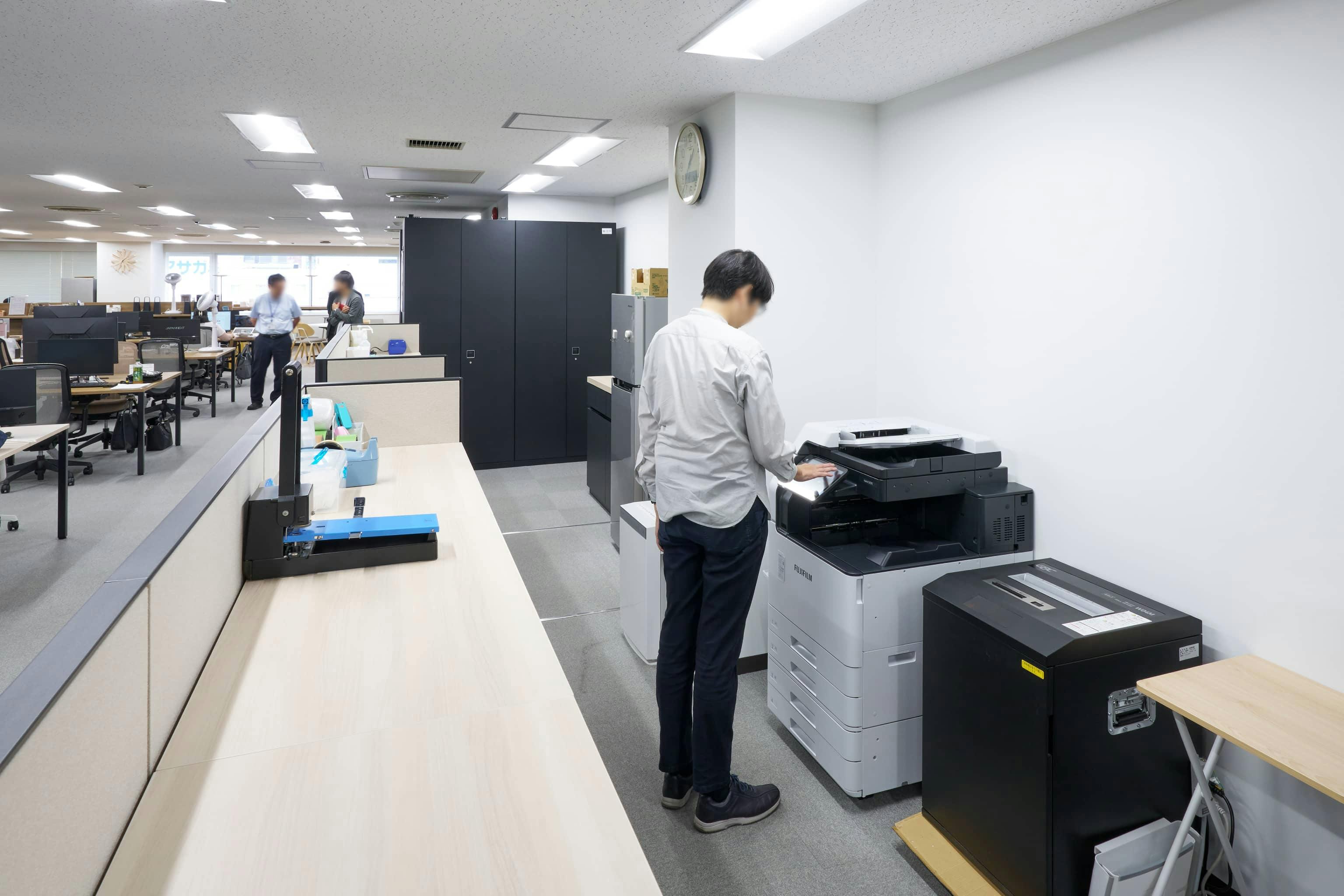 ジェイエスフィット株式会社 様の事例写真／[8F執務エリア]壁面に沿って複合機と作業台が並ぶ、限られたスペースを有効活用したレイアウト。出力した書類の整理や確認など、ちょっとした作業がしやすいよう工夫されています。作業しながら言葉を交わす社員の姿も見られます。
