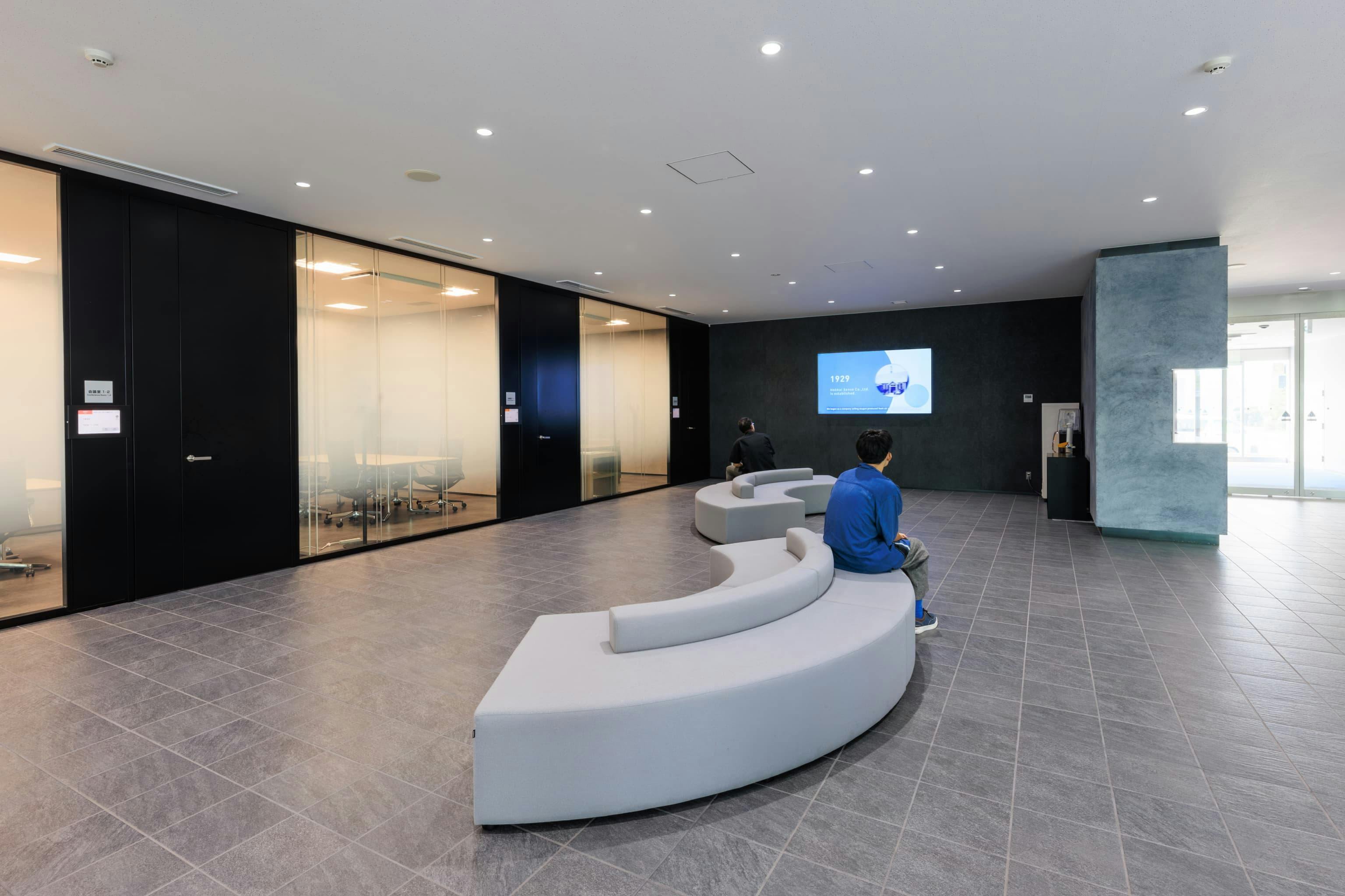 エア・ウォーター・エンジニアリング株式会社 様の事例写真／[1Fエントランスホール]カーブのついたソファが整然とした空間の遊び心あるアクセントに。両側に座ることができ、団体客にも窮屈な思いをさせない心遣いが見られます。海外からの視察を想定し、デジタルサイネージでは最新の企業情報を英語でも発信します。