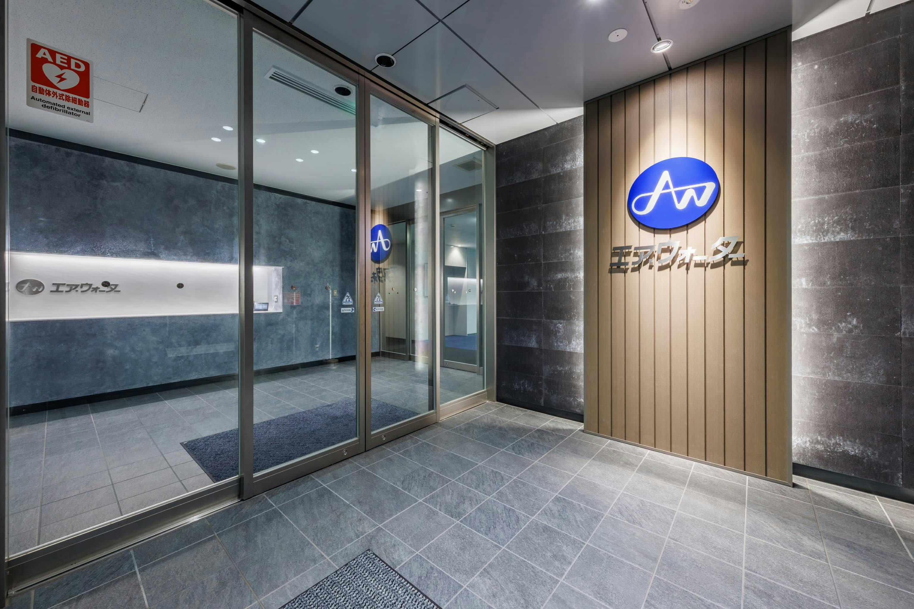 エア・ウォーター・エンジニアリング株式会社 様の事例写真／[1Fエントランス]エントランスに掲げるカンパニーサインが、エア・ウォーターグループとしての誇りを来訪者にアピール。背後のルーバーには、グループ会社が扱っている廃材を用いた素材を取り入れ、環境に配慮する姿勢を伝えます。