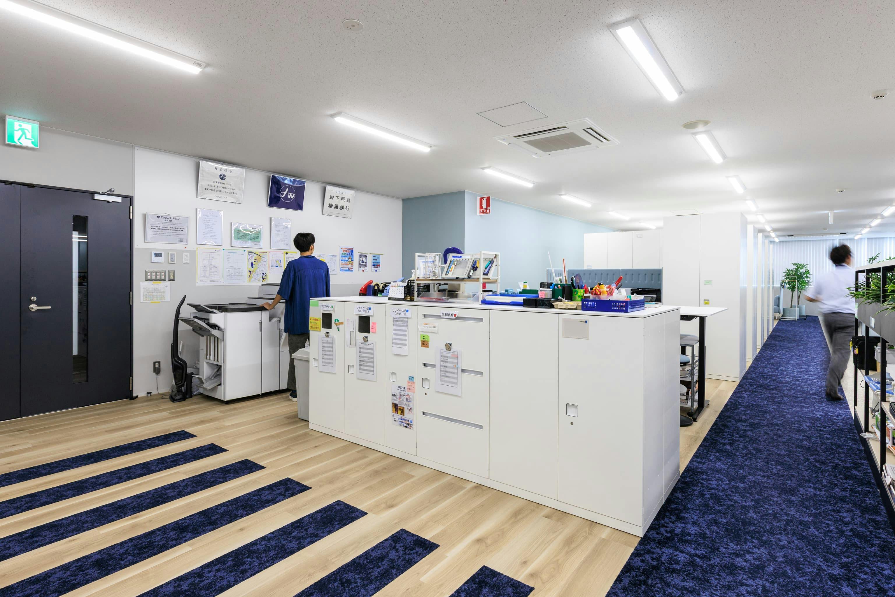 エア・ウォーター・エンジニアリング株式会社 様の事例写真／[3F執務エリア]コーポレートカラーに合わせたブルーのカーペットと板材の貼り分けにより、壁で区切ることなくゾーニングを明確化。出入り口のドアに通じる動線は横断歩道を思わせるデザインで、通行をスムーズにしています。