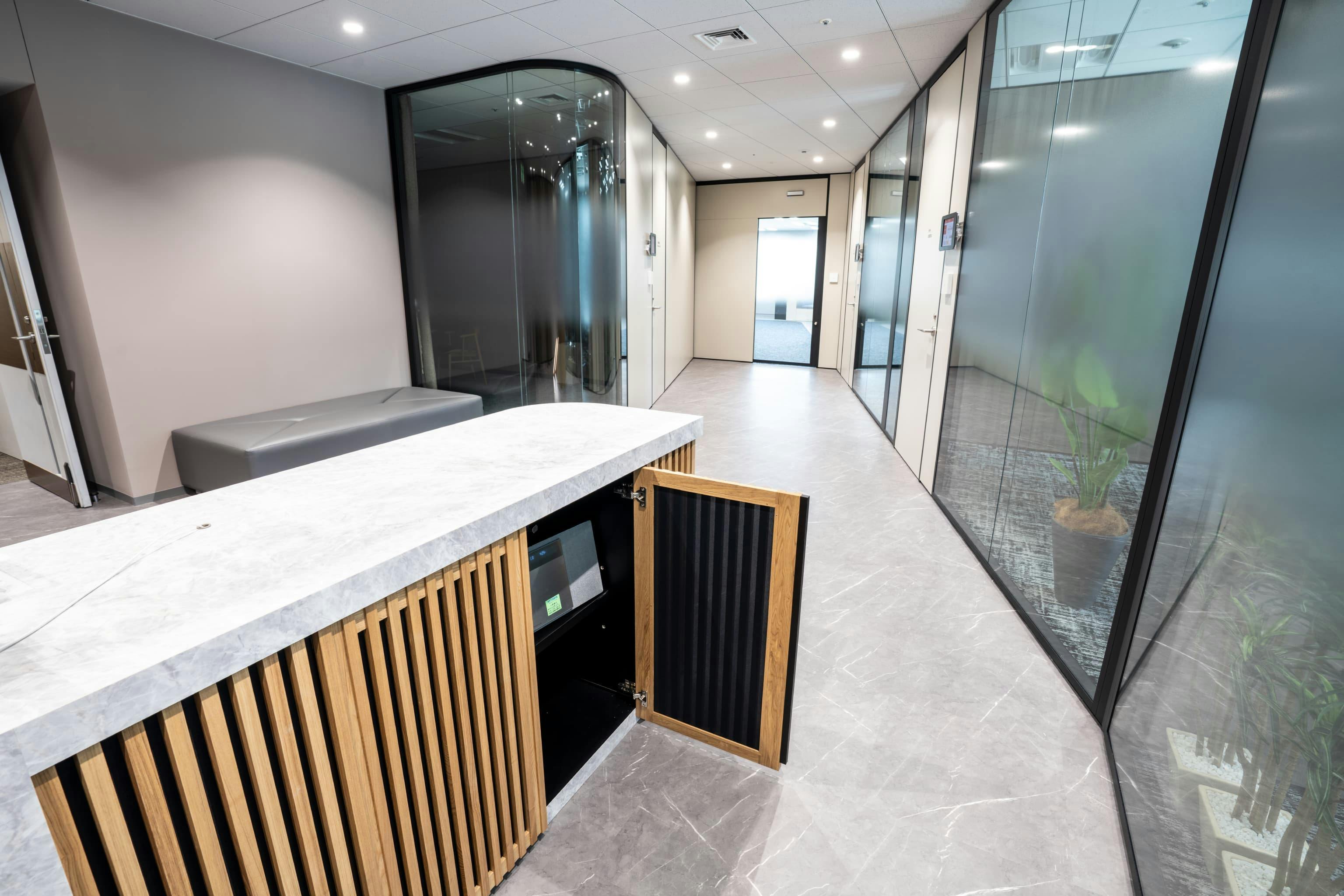 株式会社NTTデータSMS 様の事例写真／[エントランス（受付台）]受付台に内蔵されたスピーカーからピアノの音色や自然音が再生され、応接室などの会話漏れをカバー。ルーバーデザインが音の広がりと排熱に配慮しつつ軽やかさを表現し、機能的でありながら美しい設えです。