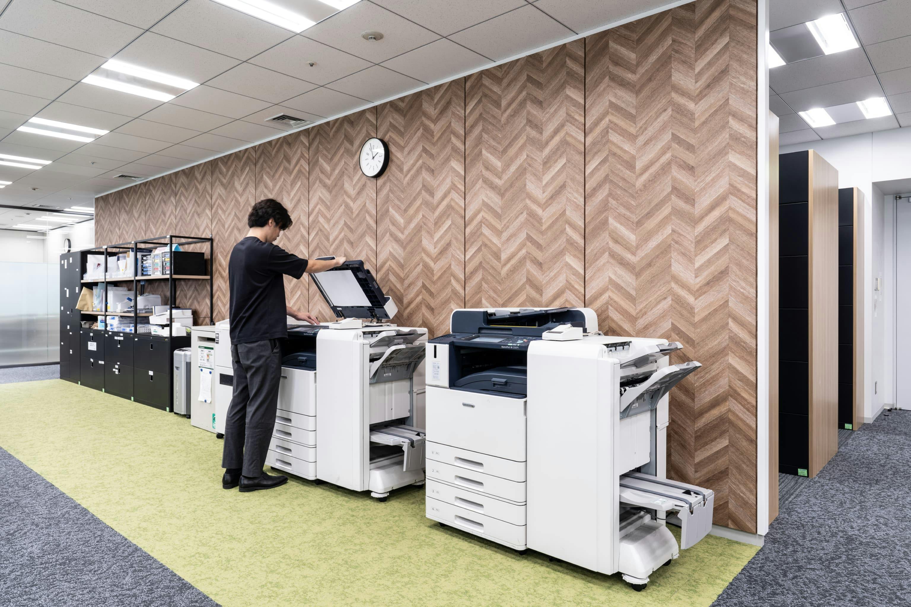 株式会社NTTデータSMS 様の事例写真／[執務エリア]コピー機などのオフィス機器は壁際の1カ所に集約され、シンプルな利用動線で事務作業が効率よく進められます。グリーンの床と木目の壁が調和し、ほっと一息つけるリラックス感が漂っています。