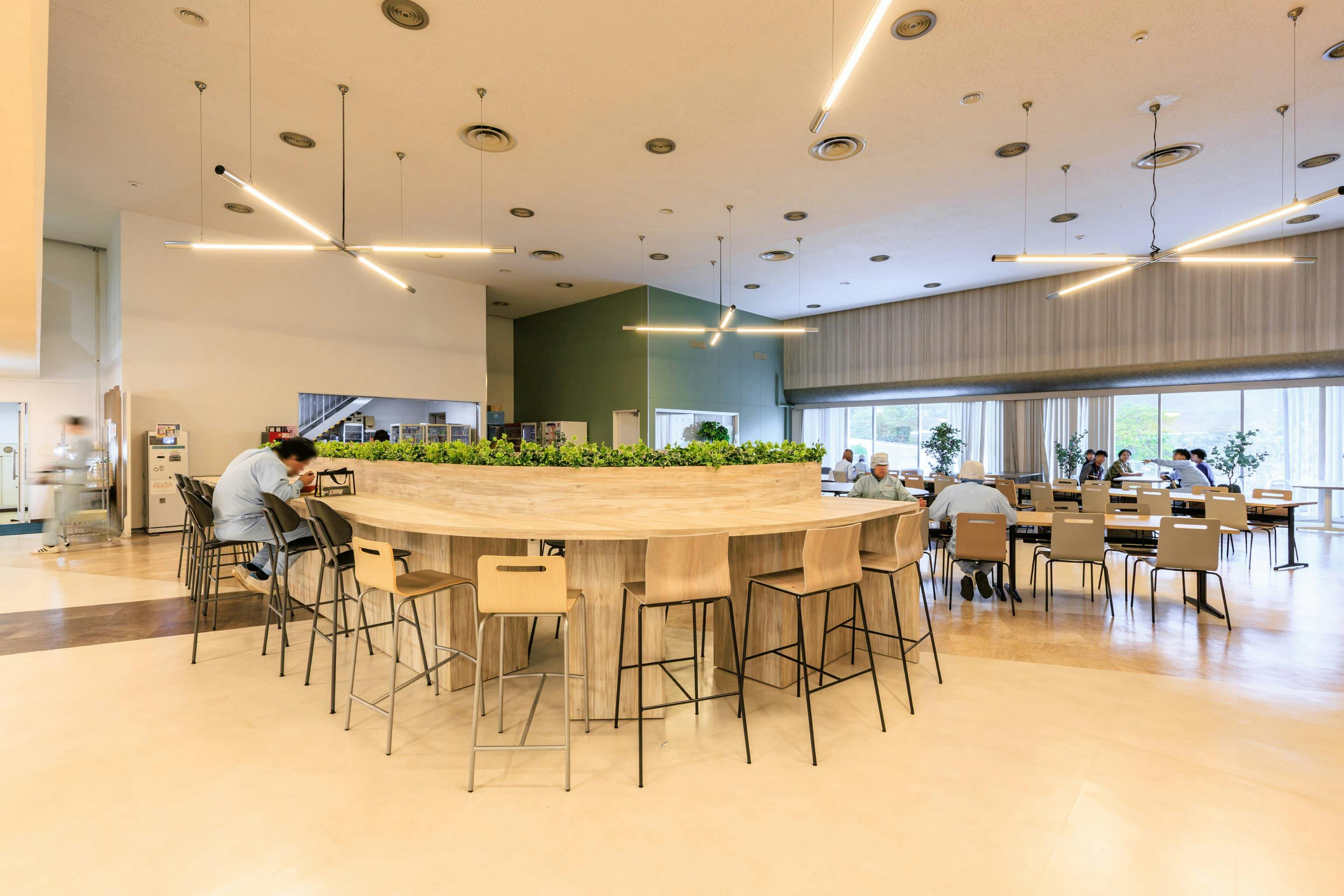 TOPPAN株式会社 ／ TOPPANパッケージプロダクツ株式会社 様の事例写真／[食堂 カウンター席]曲線を描く木の大型テーブルは、手早く食べ終えたい人にぴったりのカウンター席。一人で食事をしたい人も、隣り合った人とちょっとした挨拶や会話を交わしやすい空間です。グリーンでさりげなく視線をさえぎり、居心地の良さを演出しています。
