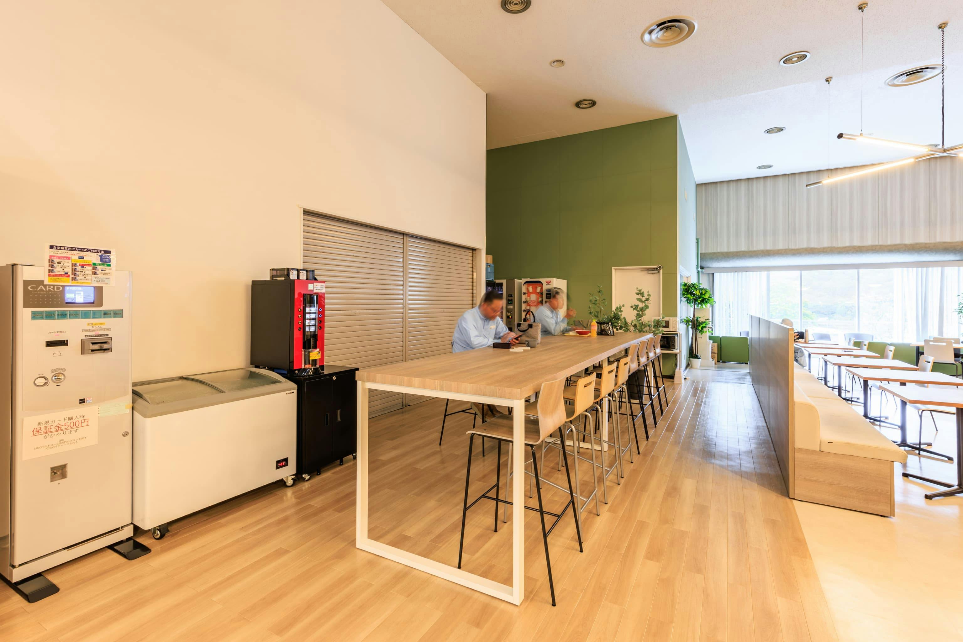 TOPPAN株式会社 ／ TOPPANパッケージプロダクツ株式会社 様の事例写真／[食堂 ハイテーブル席]軽食やドリンクを販売する売店の前には、購入したものをさっとつまめるハイテーブルを設置。写真右のソファ席の高い背もたれが、交流エリアとの自然な間仕切りになっています。