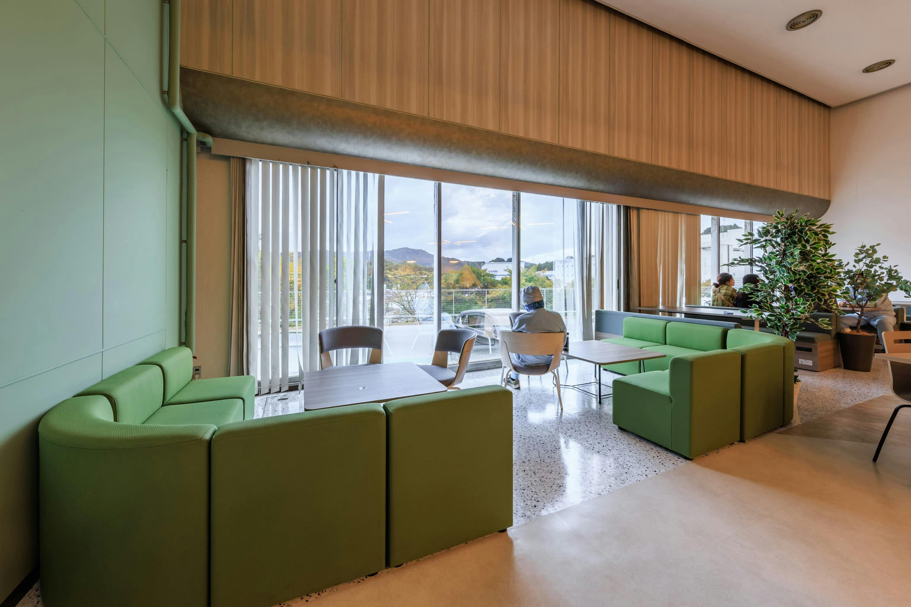 TOPPAN株式会社 ／ TOPPANパッケージプロダクツ株式会社 様の事例写真／[食堂 窓際ソファ席]窓側のソファ席やファミレス席は、一人でくつろぎたい人やじっくり話したいグループに人気で、打ち合わせにも使われています。ソファの貼り地は、外の緑に合うグリーンをチョイス。おこもり感もある落ち着いた雰囲気です。