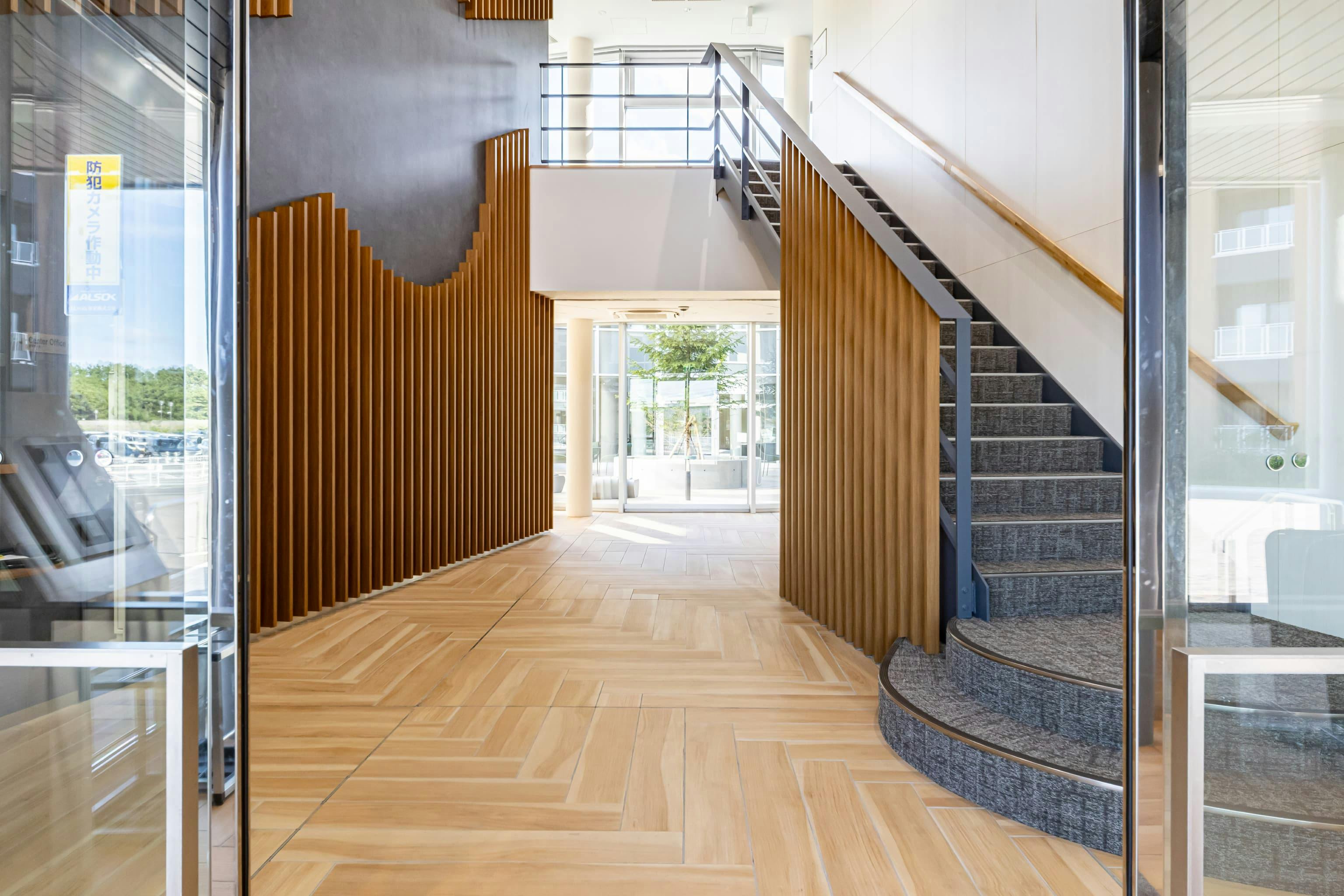 日産自動車九州株式会社 様の事例写真／[共用棟 1Fエントランス]ガラス張りで明るく広々としたエントランス。壁面のルーバーは、日産自動車のロゴマークを模した円形のデザインが施され、企業のアイデンティティをさりげなく感じさせます。