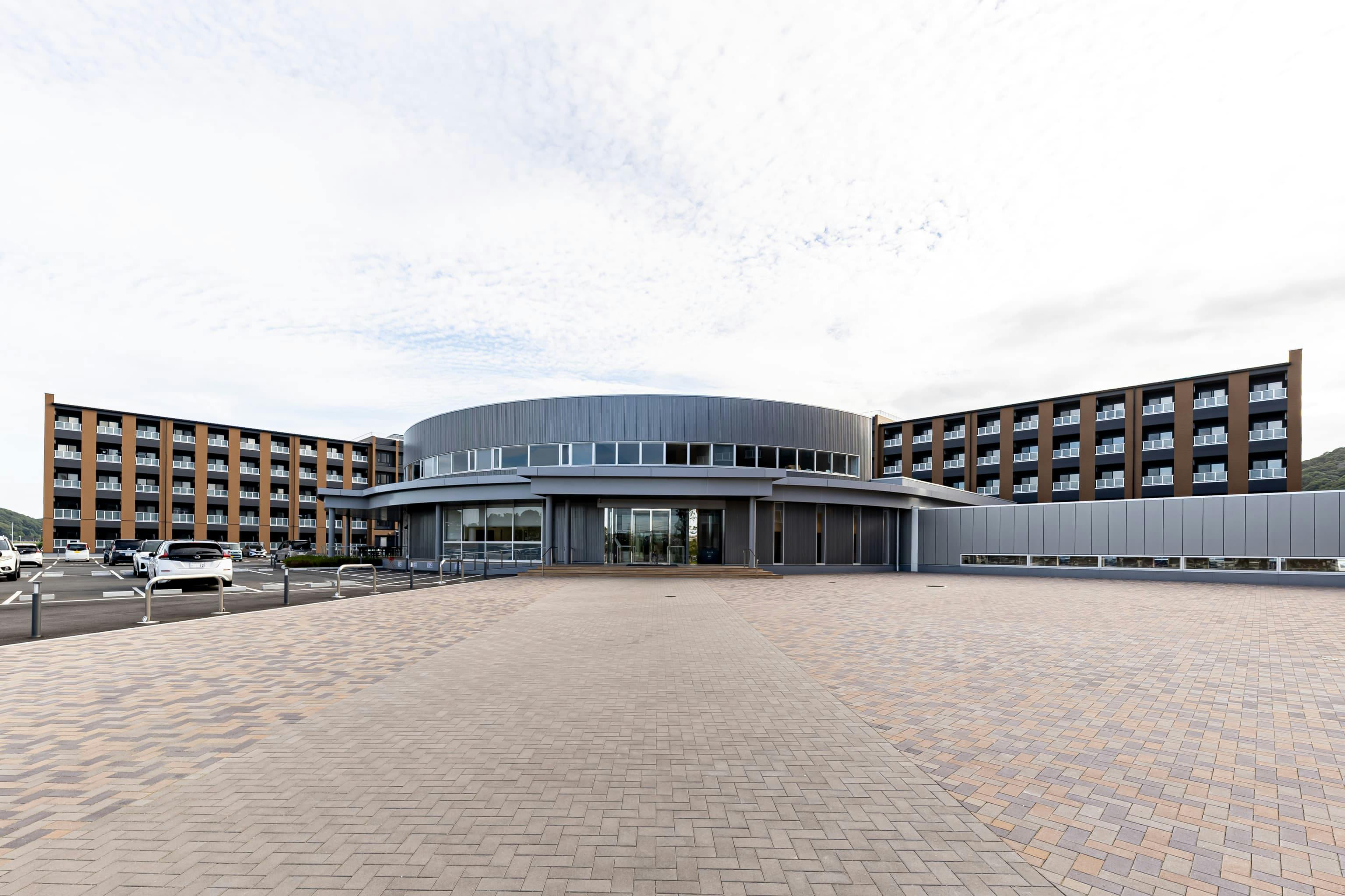 日産自動車九州株式会社 様の事例写真／[ラ・ポーズ白石 外観]中央の円形の建物が共用棟、その周辺を居住スペースであるドミトリー棟がコの字型に取り囲んでいます。共用棟と各ドミトリー棟は連絡通路でつながっており、夜間や悪天候でも安全に行き来することが可能です。