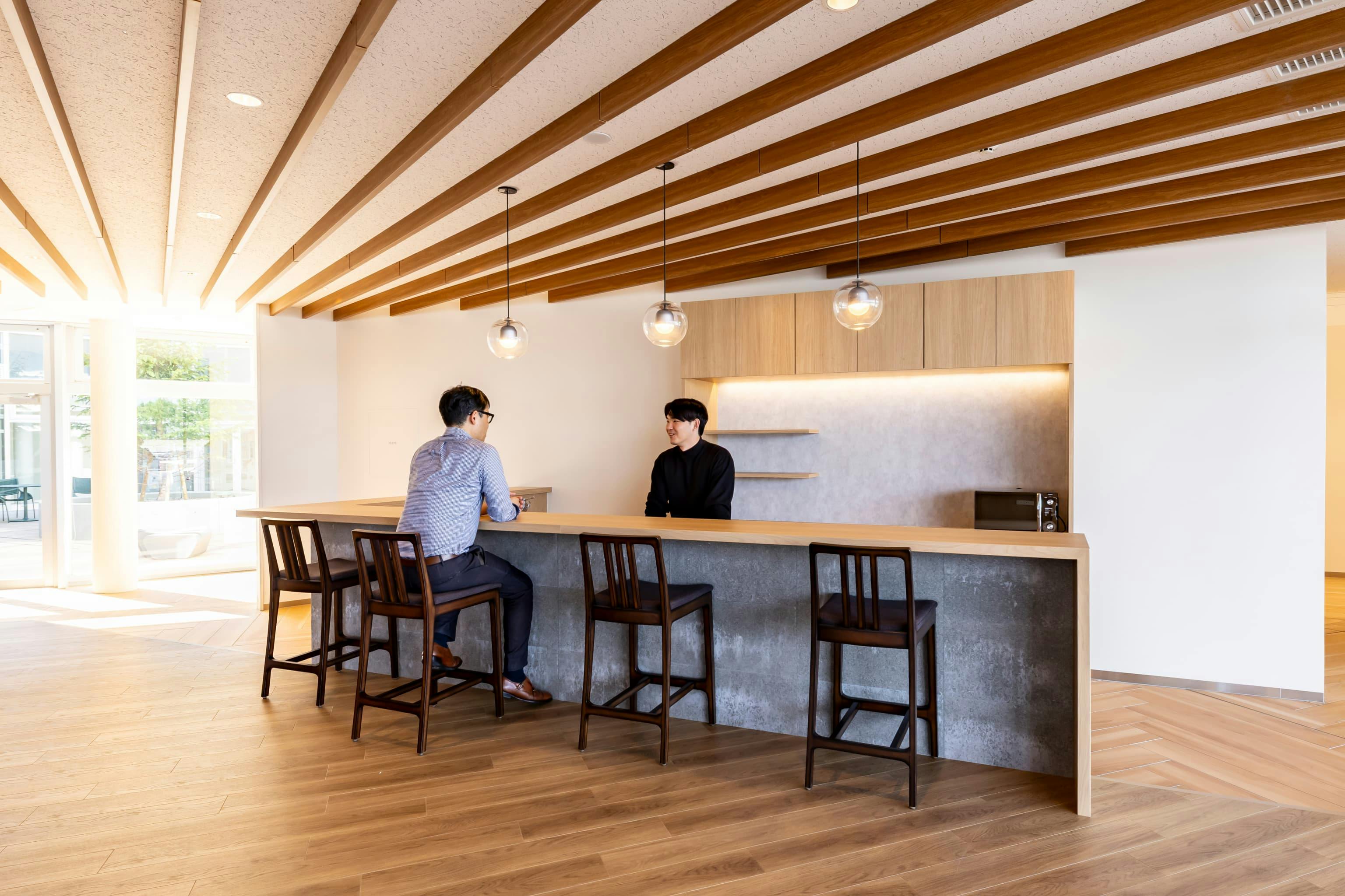 日産自動車九州株式会社 様の事例写真／[共用棟 1F コミュニティスペース]カフェカウンターは出勤前にコーヒーを飲んだり、就業後に食事やお酒を楽しんだりと、さまざまな過ごし方ができる場所。小上がりの段差を活かし、カウンター越しに座る人と立つ人の目線が合うよう設計されています。
