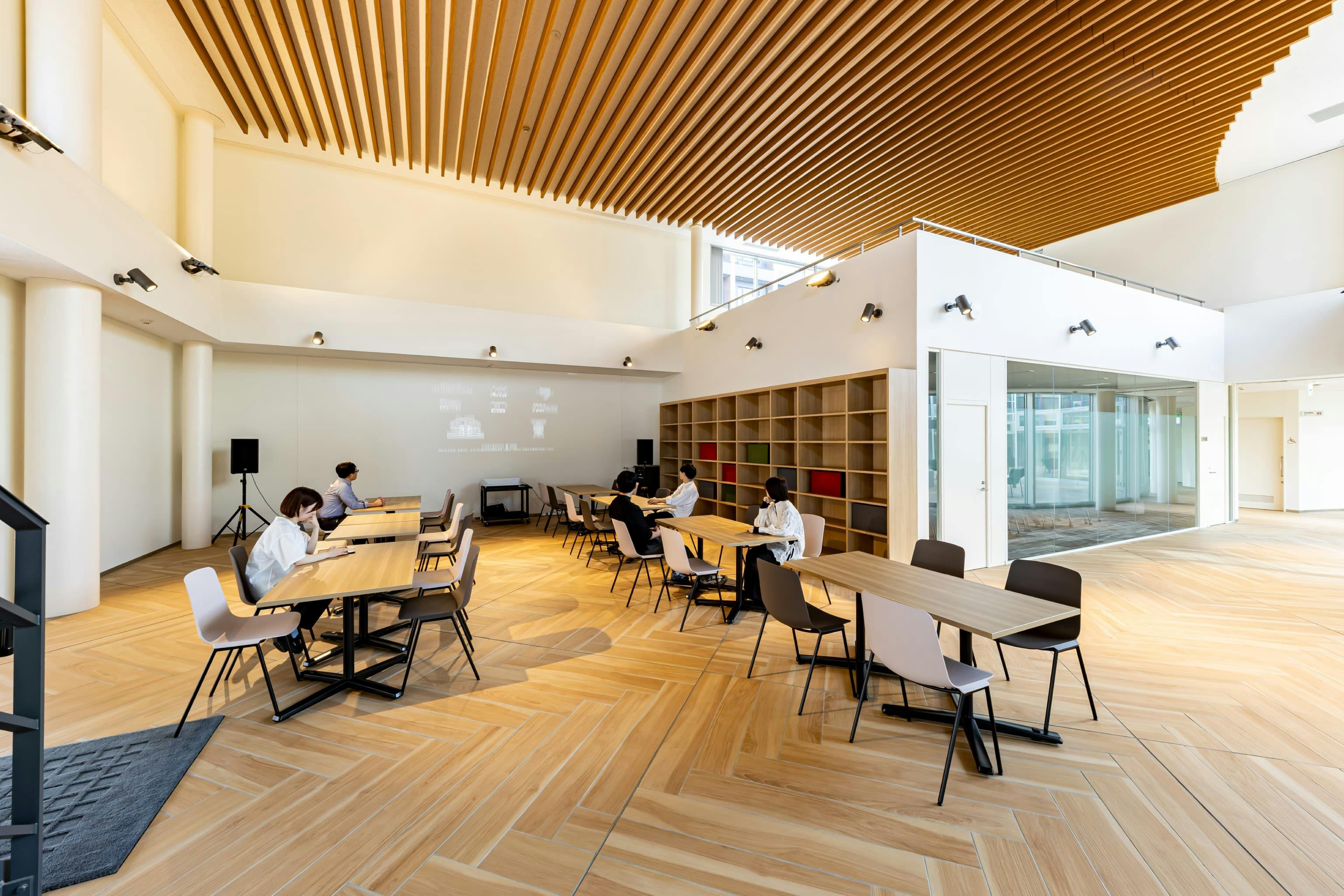 日産自動車九州株式会社 様の事例写真／[共用棟 1F ビューイングスペース]柔軟にレイアウトを変更できる可動式のテーブルとチェアを用意。みんなで遊ぶボードゲームや本などを置けるよう、壁面に木製棚が設けられています。AV機器は更新や入れ替えがしやすい据え置きタイプです。