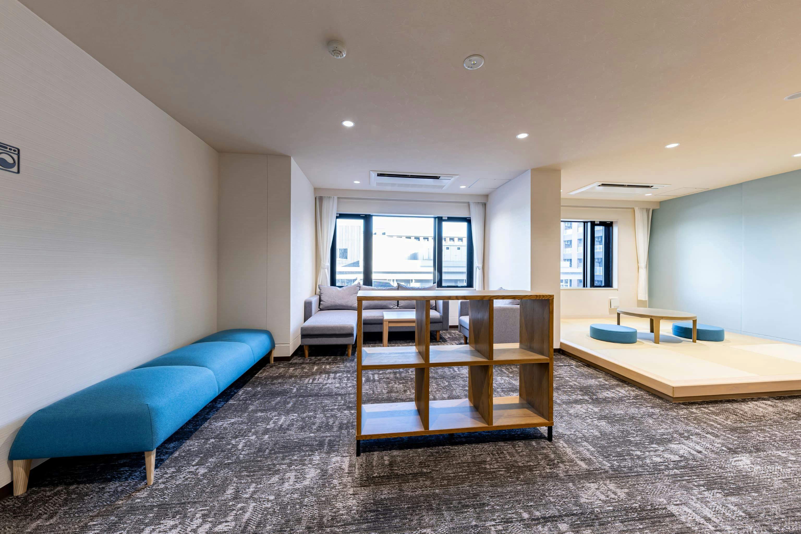 日産自動車九州株式会社 様の事例写真／[ドミトリー棟 3Fラウンジ]その日の気分で過ごすフロアを選べるよう、「海の青」「木々の緑」「夕焼けのオレンジ」など自然を意識したカラーを採用。中央の棚がキッチンへの動線とランドリーからソファへ向かう動線を緩やかに分けています。