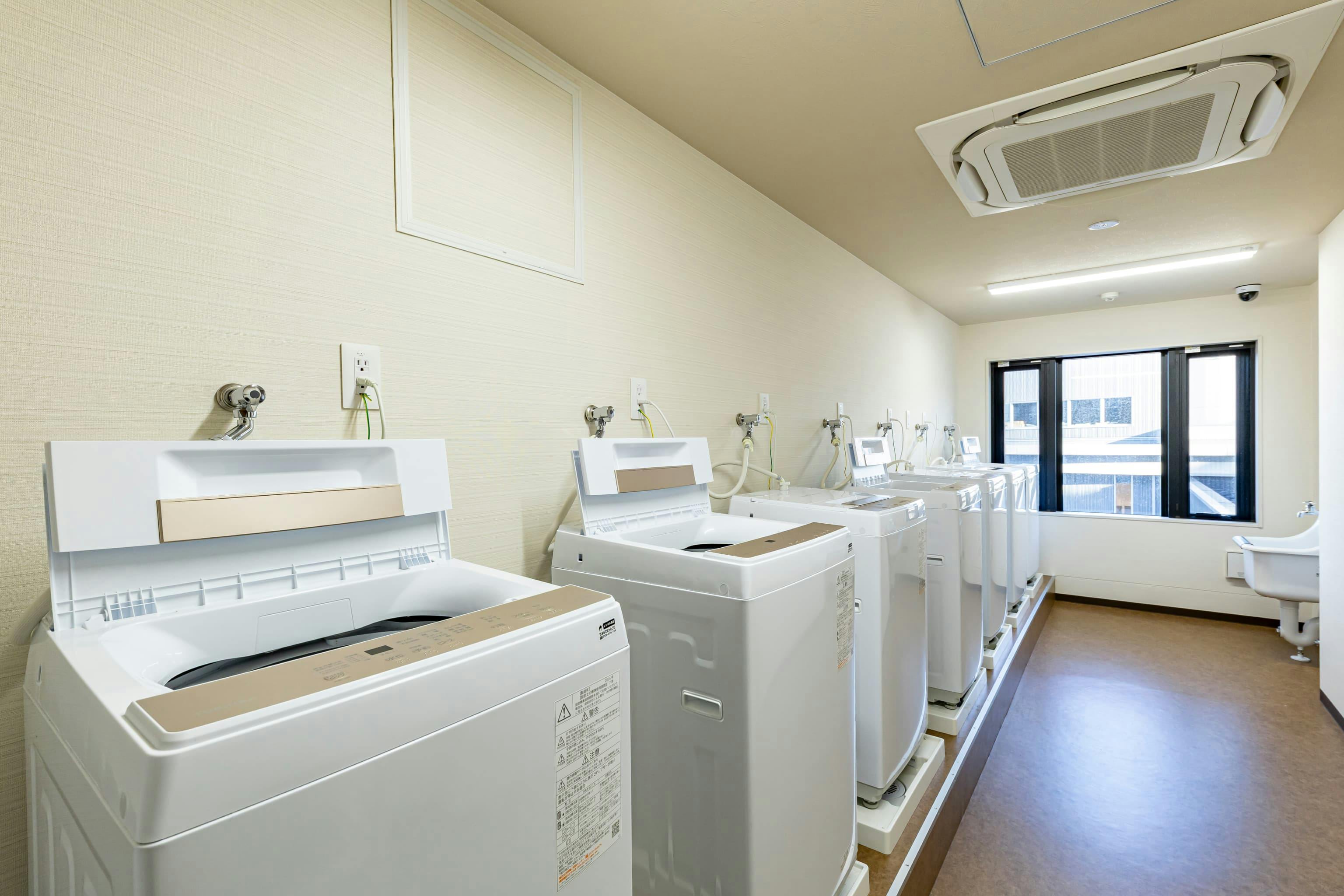 日産自動車九州株式会社 様の事例写真／[ドミトリー棟 ランドリールーム]居室内に洗濯機置き場を設けない代わりに、各階には複数台の洗濯機や乾燥機を設置したランドリールームが用意されています。ラウンジに併設することで、待ち時間もくつろぎながら過ごせます。