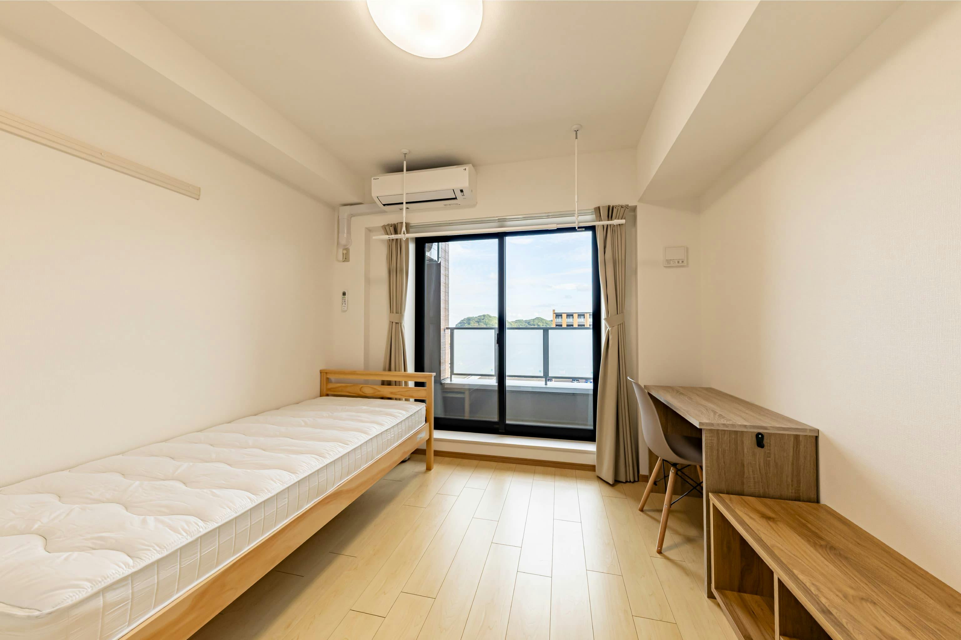 日産自動車九州株式会社 様の事例写真／[ドミトリー棟 居室]居室は約17㎡のシンプルなワンルーム。ミニキッチン、浴室、トイレを備え、ベッドやデスク、ローボードなど生活に必要な家具があらかじめ用意されています。必要最低限の私物を持ち込むだけで、新生活を始められる環境です。