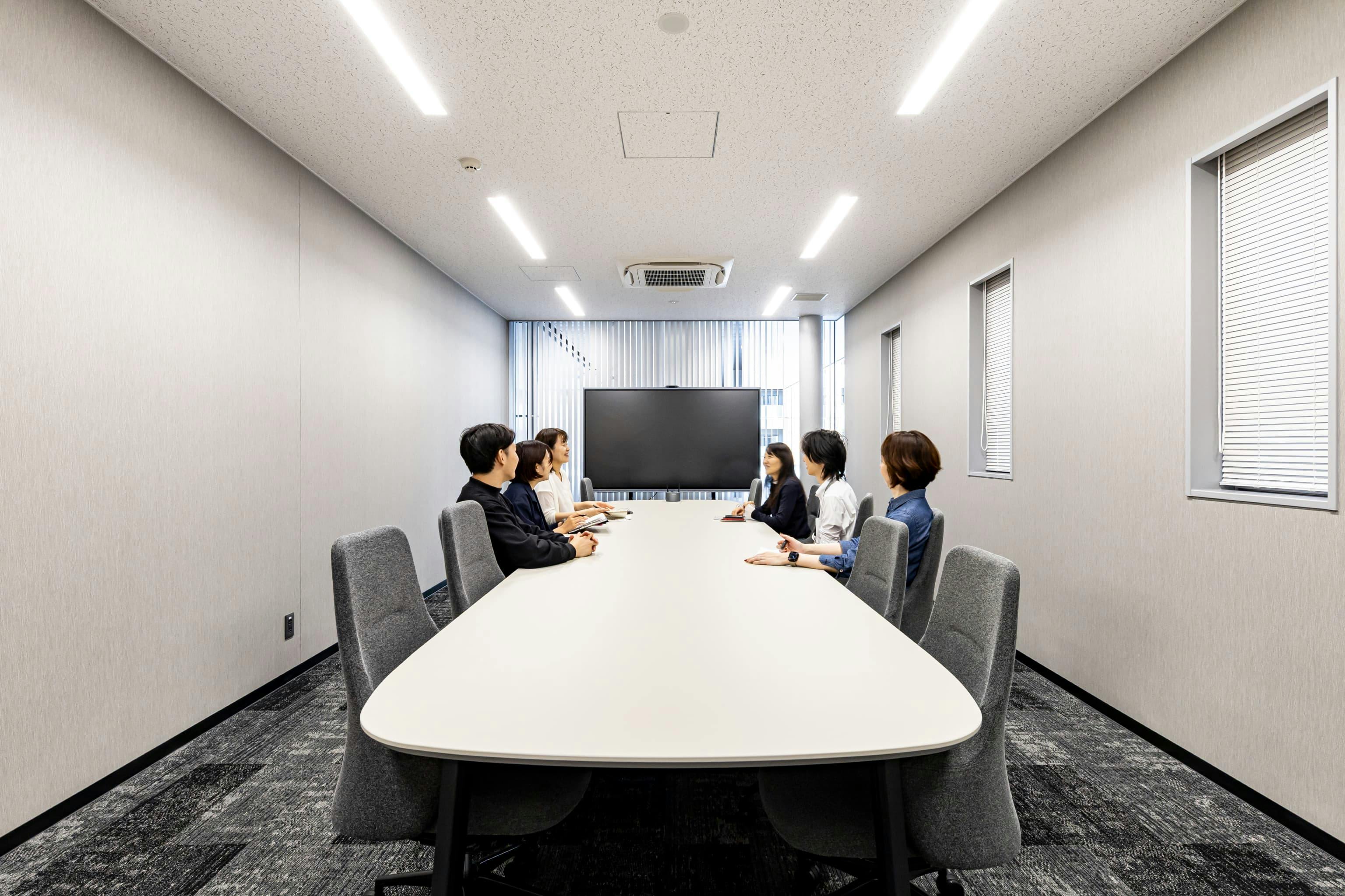 日産自動車九州株式会社 様の事例写真／[共用棟 1F 会議室「ELGRAND」]応接室を兼ねる会議室「ELGRAND」は、役員来訪時や家族との面談など、クローズドな話をしたい場面にも適した空間です。落ち着いたグレー基調の内装と重厚感のある設えが、安心して話せる雰囲気を演出しています。