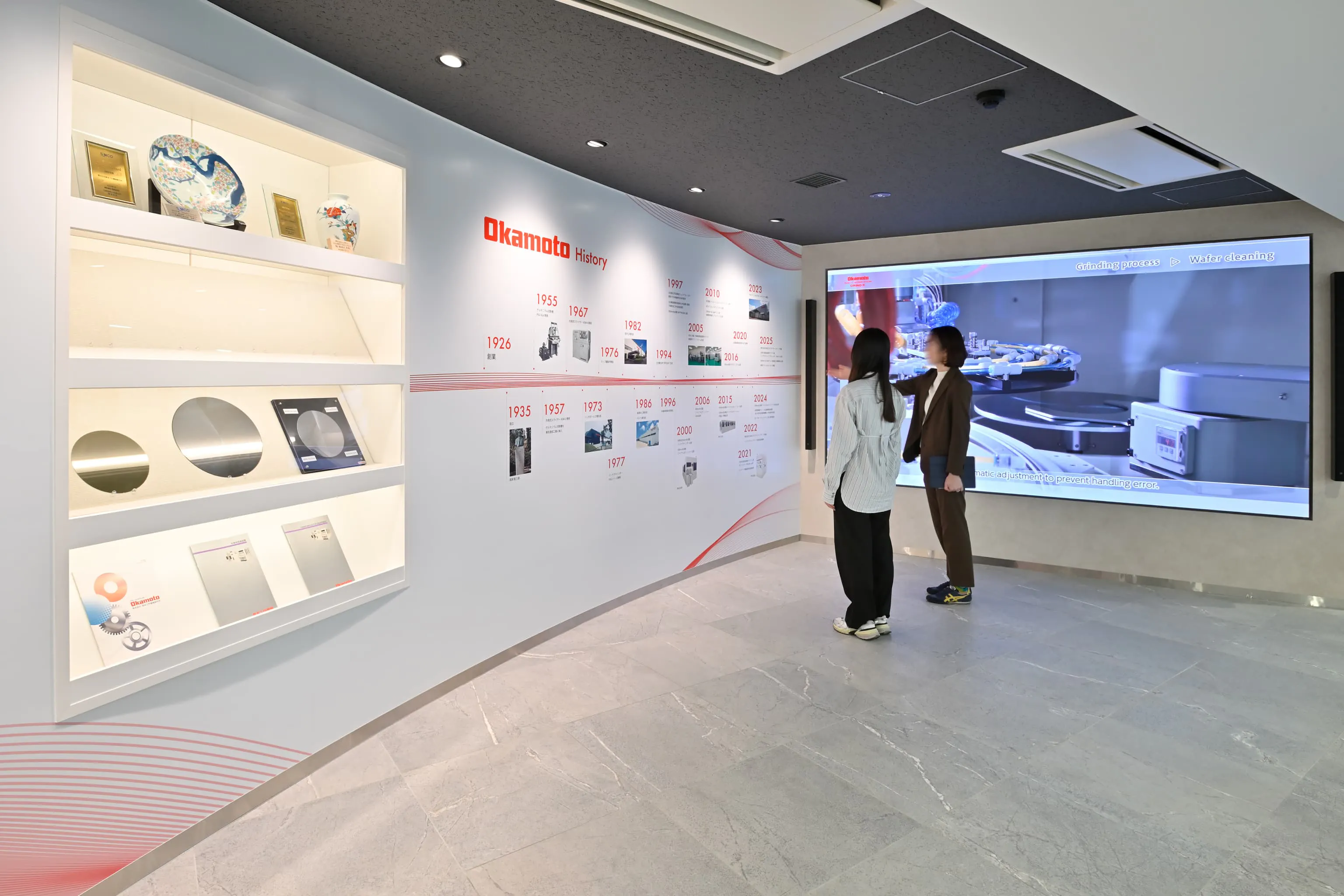 株式会社岡本工作機械製作所 様の事例写真／[1F PRスペース]大型モニターや壁面棚を活用し、企業の歴史や技術背景を伝えるPRスペースです。展示棚は入れ替えやすく、製品サンプルや最新トピックを柔軟に紹介できます。ショールーム見学の前に企業を知ってもらう重要な場です。