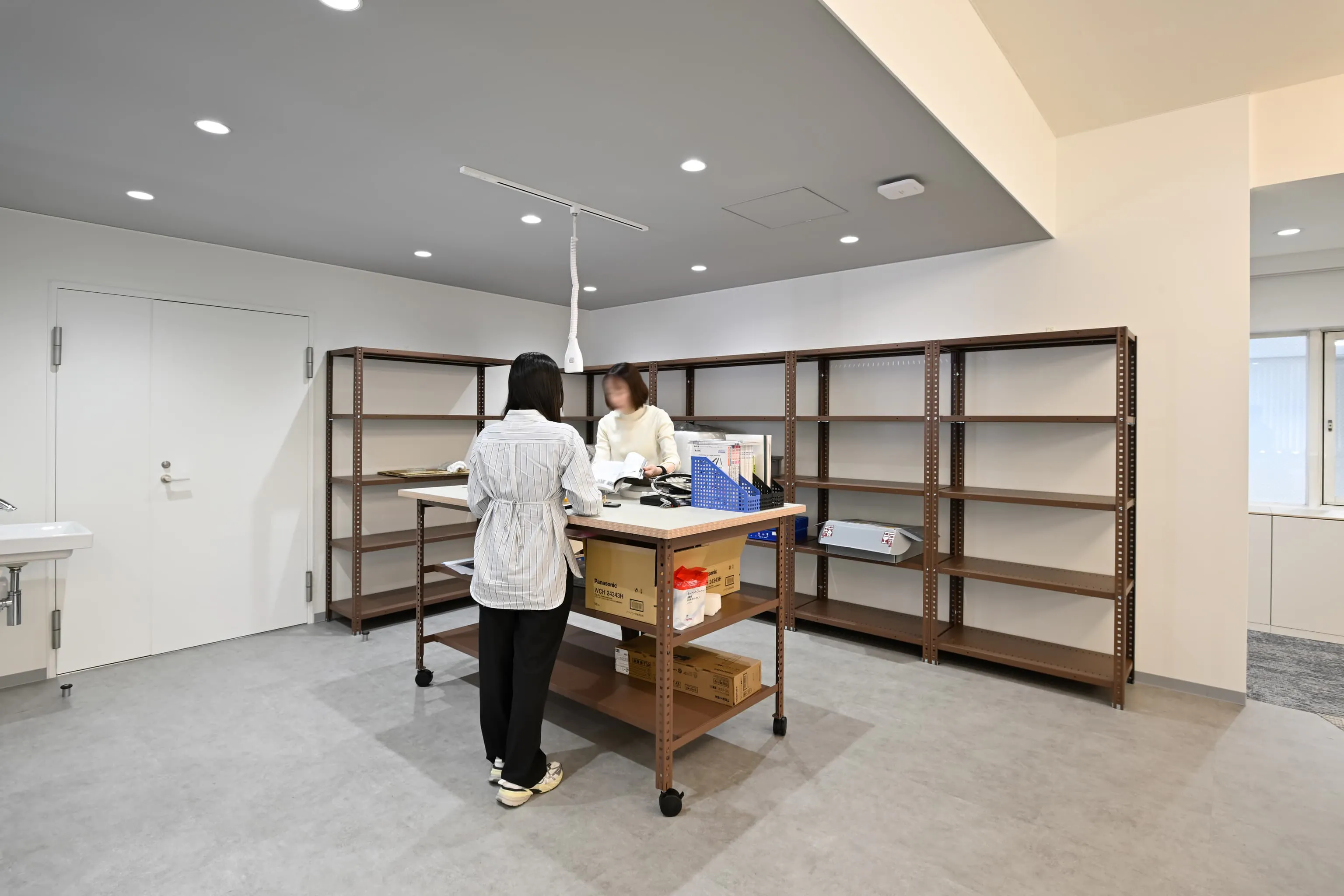 株式会社岡本工作機械製作所 様の事例写真／[2F作業スペース]厨房だった場所を大幅に改装して設置された作業スペース。大きな作業台では製品模型やパーツを手に取りながら開発を進められます。モルタル調のグレー床とブラウン系スチール棚が工房のような雰囲気を演出しています。