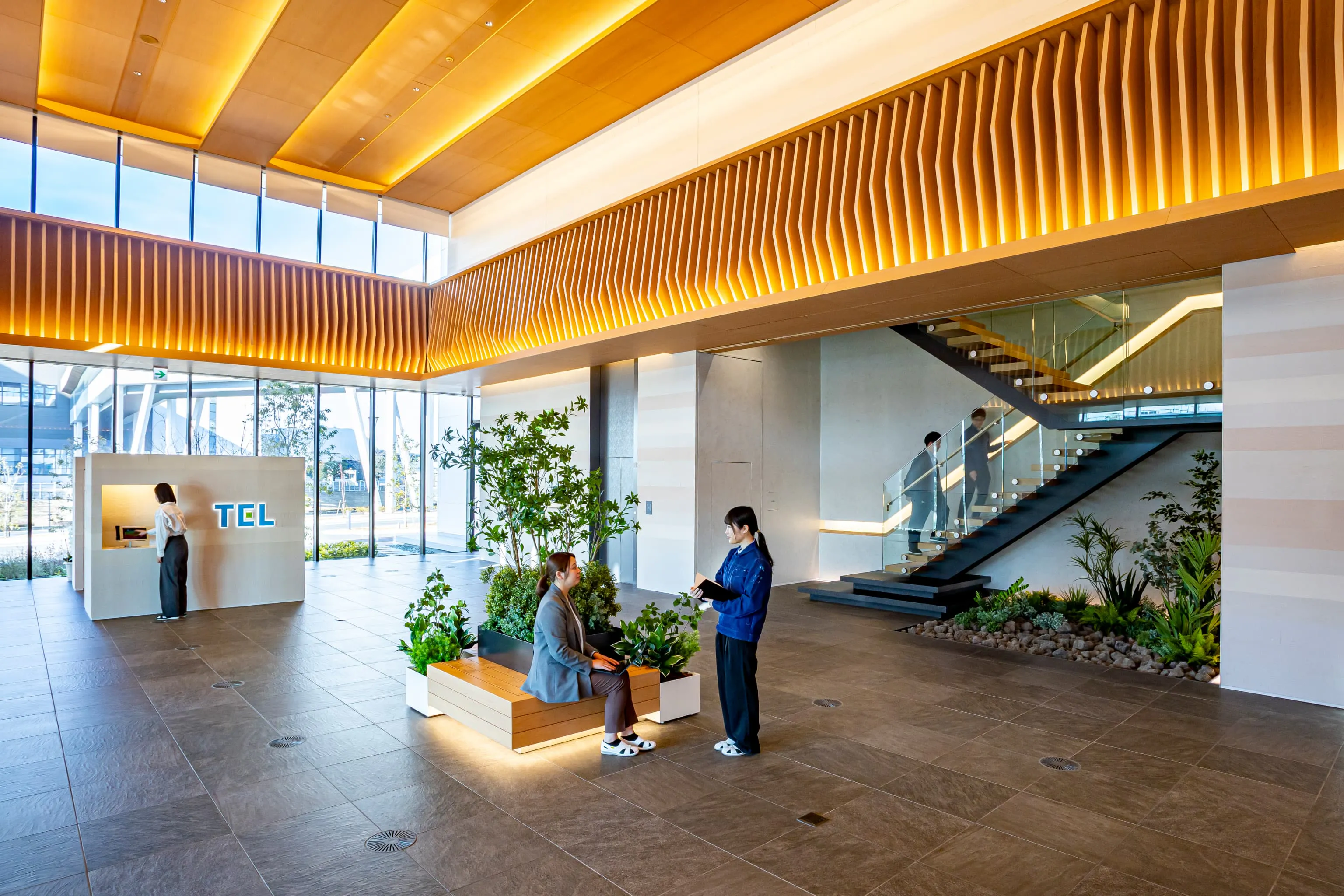 東京エレクトロン九州株式会社 様の事例写真／[1Fエントランス]企業ロゴが目を引く受付台は、熊本の土を使用した塗り壁です。階段下の植栽エリアには阿蘇山の溶岩石が敷かれ、壁面はさまざまな色が重なり阿蘇の地層が表現されています。来訪者に熊本の地域性を感じさせる空間です。
