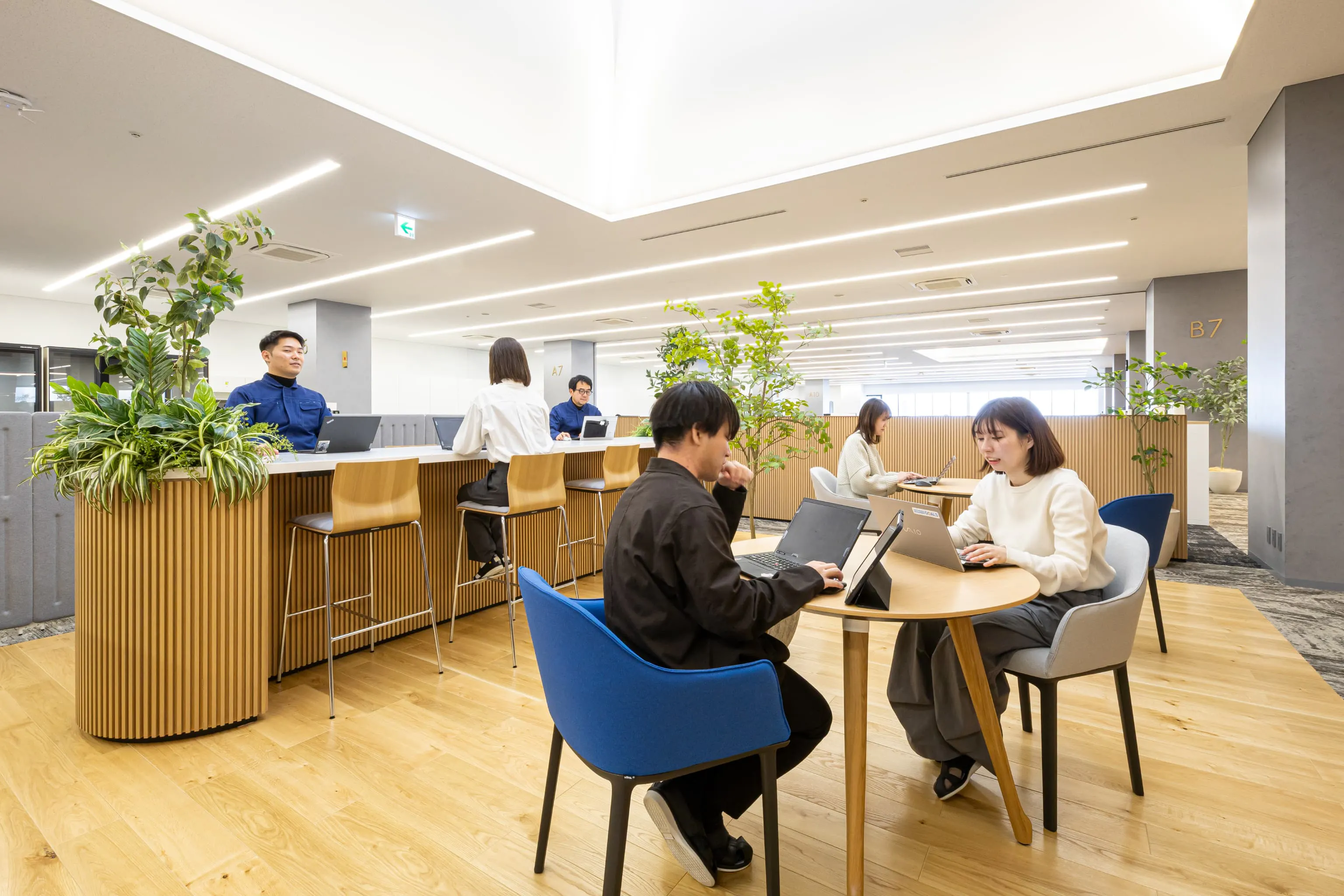東京エレクトロン九州株式会社 様の事例写真／[4F執務エリア]こちらのトップライト下には、打ち合わせや集中作業など多目的に使えるカフェ形式の席が配置されています。木のパーティションが周囲からの視線を緩やかに遮ることで、開放感と適度なこもり感を両立した空間を作り出しています。