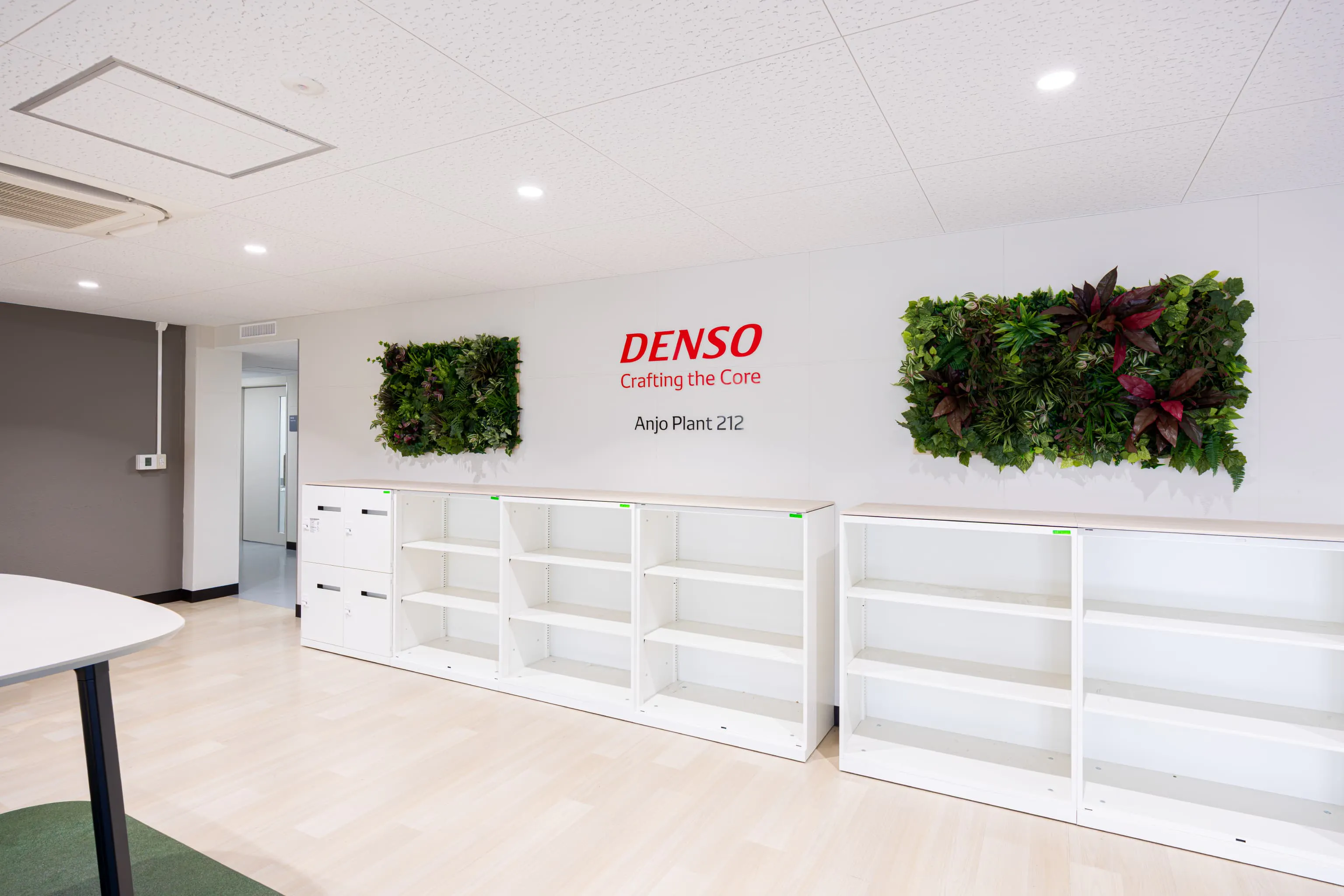 株式会社デンソー安城製作所 様の事例写真／[1F多目的ロビー]壁面には企業ロゴ、左右には癒しをもたらす植栽が設置されています。白い棚は、郵便物や宅配物の置き場として活用され、配送業者との連携がスムーズです。