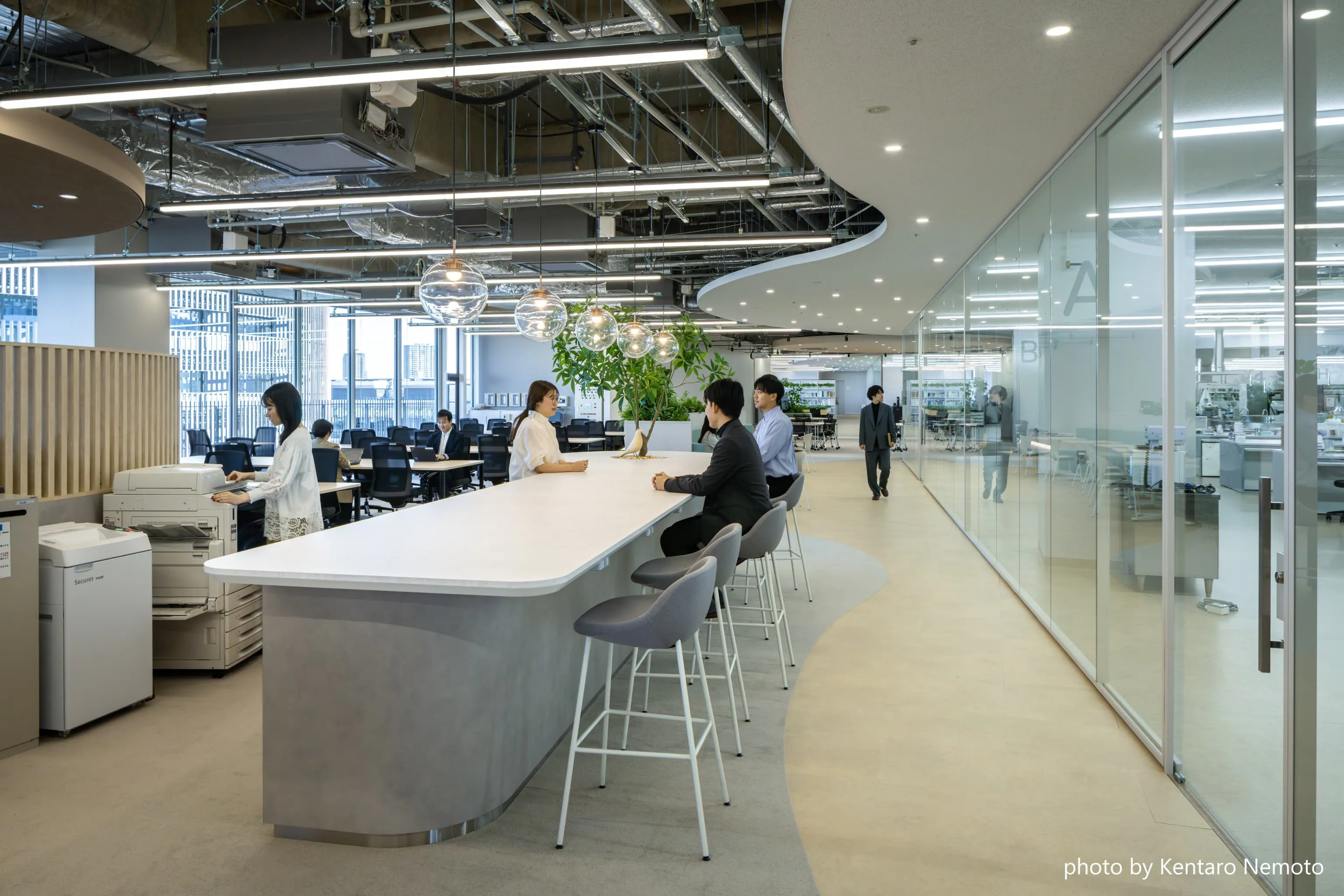 アサヒグループ食品株式会社 様の事例写真／[マグネットエリア]複合機や文具、試作品などが置かれている共有カウンターは、自然と人が引き寄せられる交流の起点です。用のついでに立ち寄る人や、カウンターで作業する人が顔を合わせ談笑する姿が見られます。