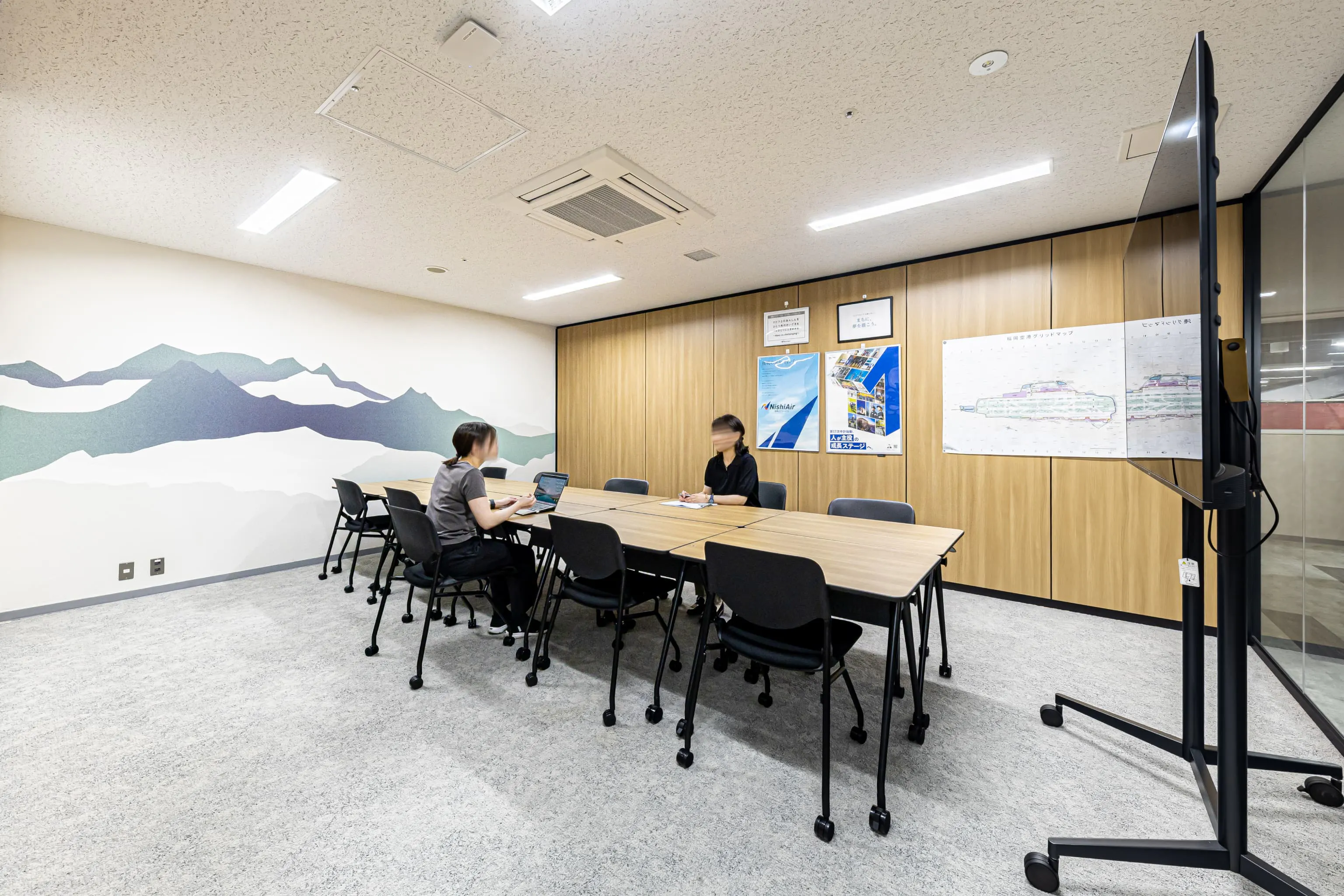 西鉄エアサービス株式会社 様の事例写真／[会議室]窓がない部屋のため、閉塞感が出ないように山のグラフィックが描かれた壁紙を採用。壁の一部をガラス張りにして抜け感が演出され、什器は研修や会議など多用途に対応できる可動式のものが選ばれています。
