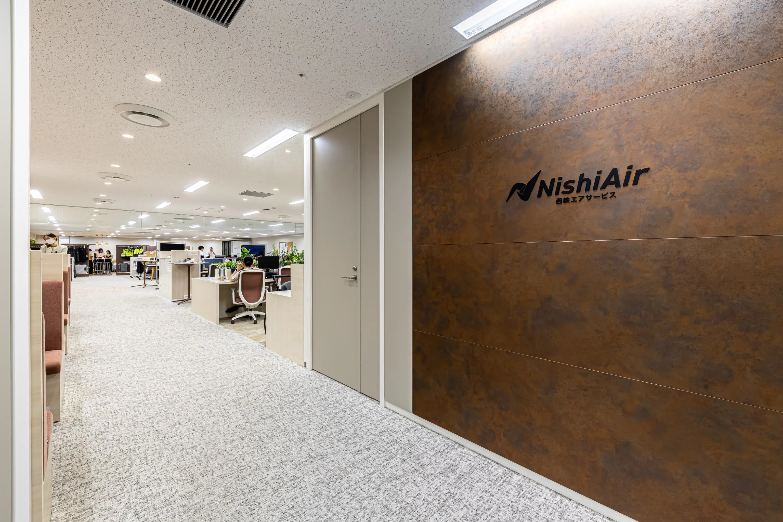 西鉄エアサービス株式会社 様の事例写真／[受付]エイジング加工が施された壁面で、スタイリッシュな佇まいに仕上げられた受付。背景の独特な質感に合わせ、あえて黒一色で統一されたクールなブランドロゴが美しく調和し、目を引きます。